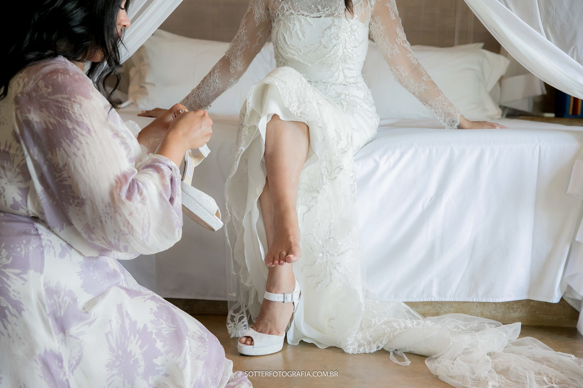 noiva sentada  na cama tendo ajuda da madrinha para calçar o seu sapato