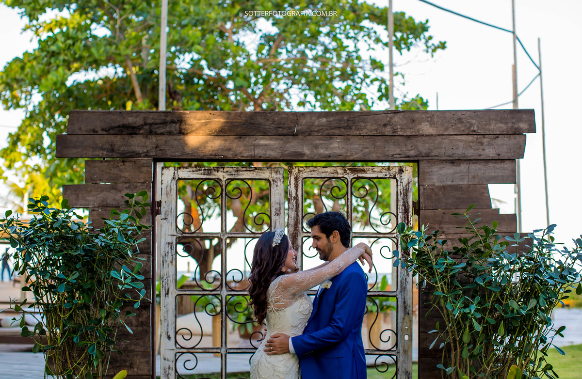 noiva com os braços apoiados nos ombros do noivo , ao fundo o portal de madeira com plantas verdes e uma porta de ferro 