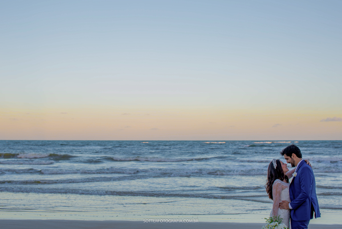 um mar azul, um céu laranja e os noivos se olhando no canto direito da imagem