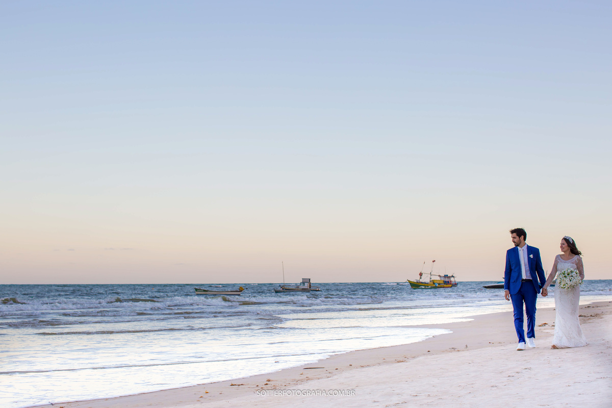 noivos caminhando na praia em trancoso, ao fundo um barco no mar deixando o cernário mais bonito, ela segura o buquê, ele usa um terno azul , foto da sotter fotografia