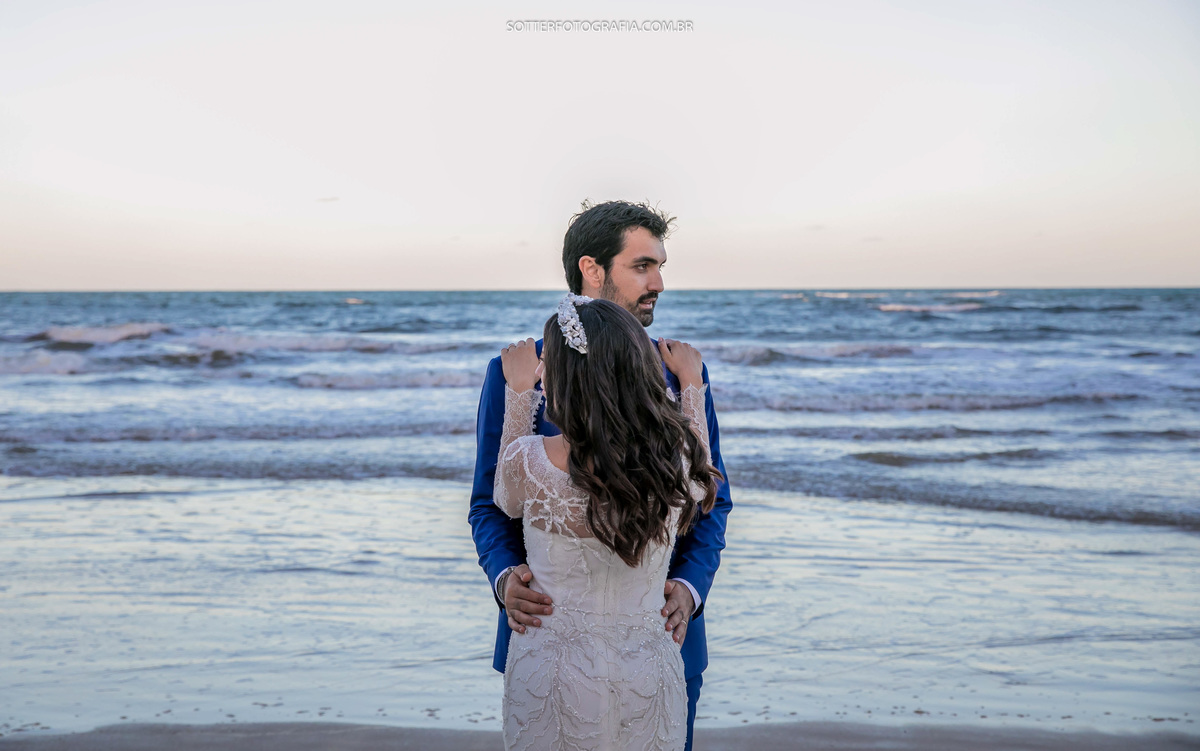 o mar ao fundo, o noivo de frente para a noiva e a noiva de costa ma imagem cada um olhando para um lado da praia
