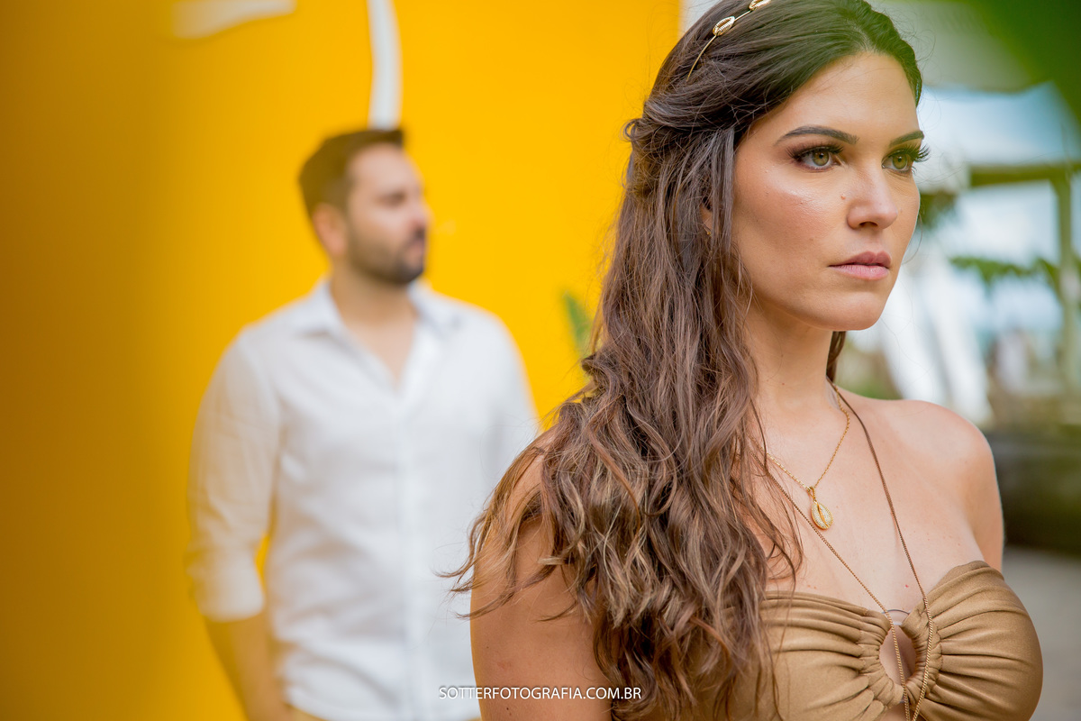ensaio pre casamento em arraial dajuda sotter fotografia noiva em destaque usando roupa dourada e noivo ao fundo usando camisa branca 