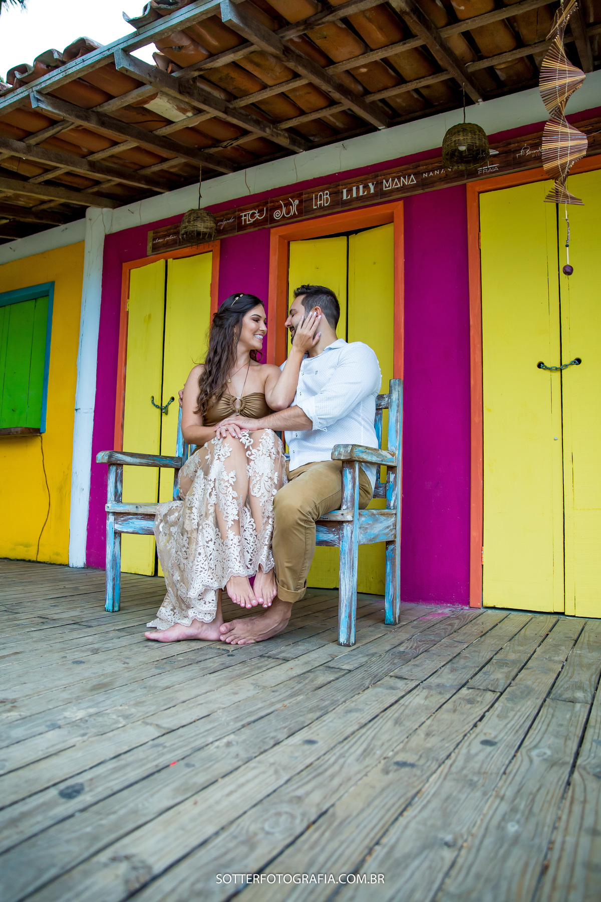 casal sentando em um banco rustico azul em frete de uma casa rosa e laranja com portas amarelas sotter fotografia arraial dajuda 