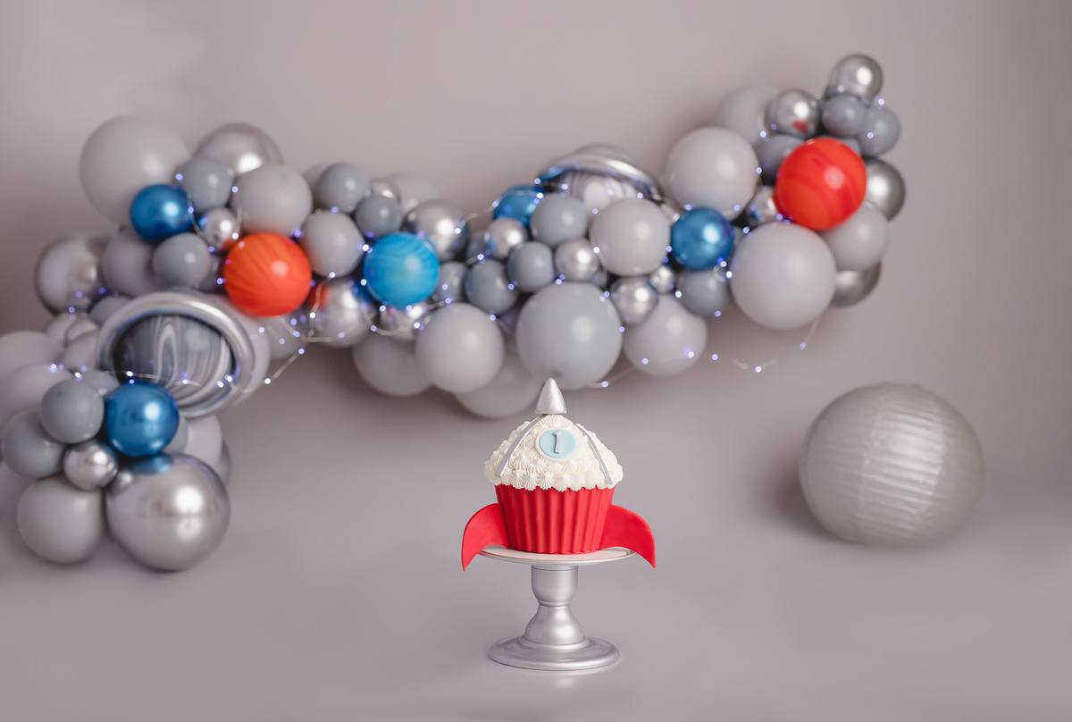 Bebe astronauta no ensaio fotografico de 1 aninho ou primeiro aninho, cenário com fundo cinza e arco de balões imitando planetas e bolo em formato de foguete vermelho com branco em cima de um suporte prata