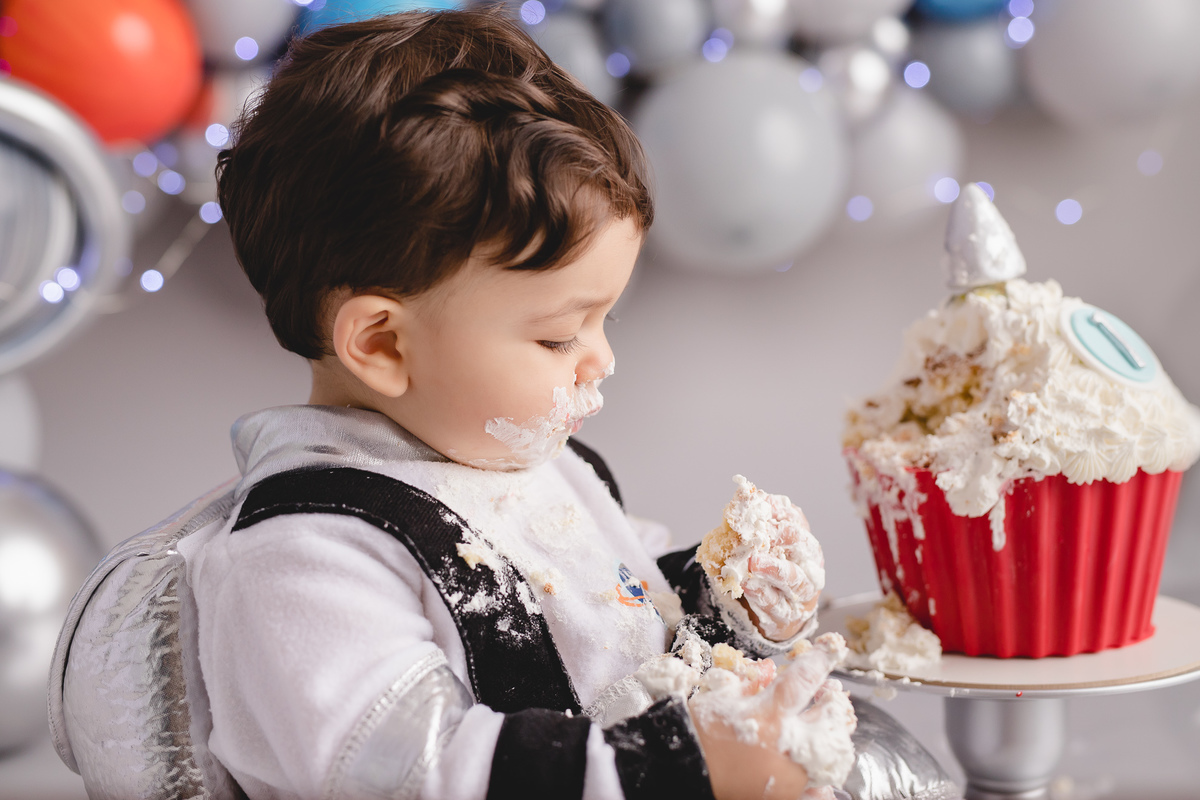 bebê astronauta no ensaio fotografico de 1 aninho ou primeiro aninho cenário cinza e arco de balões imitando planetas bolo em formato de foguete vermelho e branco em um suporte prata, detalhe bebê comendo um pedaço do bolo