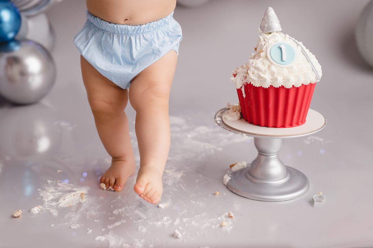planetas bolo em formato de foguete vermelho e branco em um suporte prata, detalhe bebê comendo um pedaço do bolo com tapa fralda azul e gravata branca detalhe dos pés