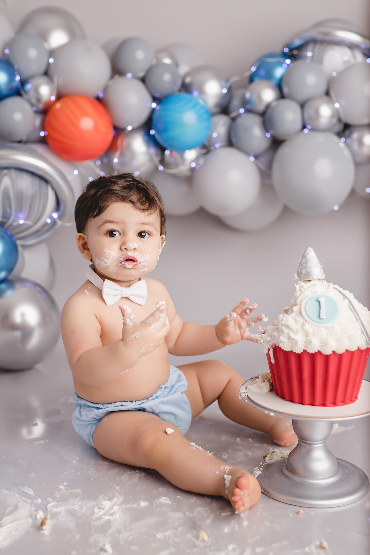 planetas bolo em formato de foguete vermelho e branco em um suporte prata, detalhe bebê comendo um pedaço do bolo com tapa fralda azul e gravata branca