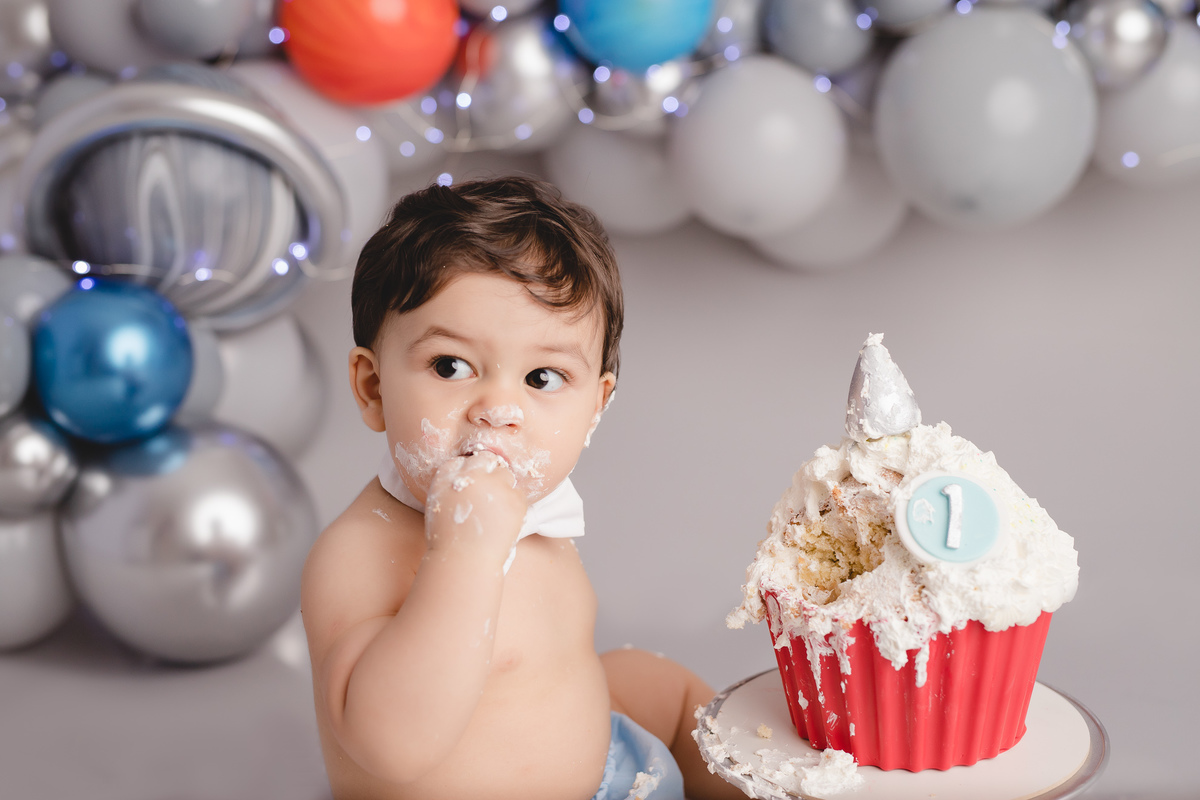 planetas bolo em formato de foguete vermelho e branco em um suporte prata, detalhe bebê comendo um pedaço do bolo com tapa fralda azul e gravata branca