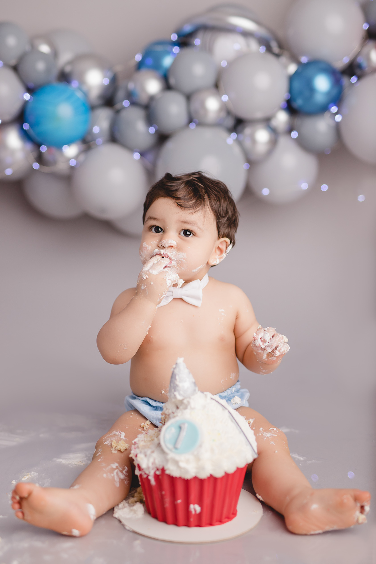 planetas bolo em formato de foguete vermelho e branco em um suporte prata, detalhe bebê comendo um pedaço do bolo com tapa fralda azul e gravata branca chupando os dedos