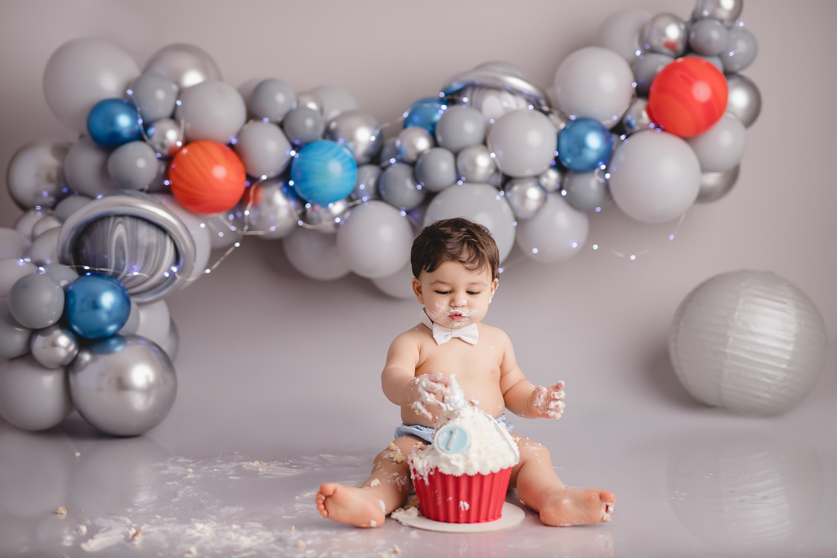 planetas bolo em formato de foguete vermelho e branco em um suporte prata, detalhe bebê comendo um pedaço do bolo com tapa fralda azul e gravata branca