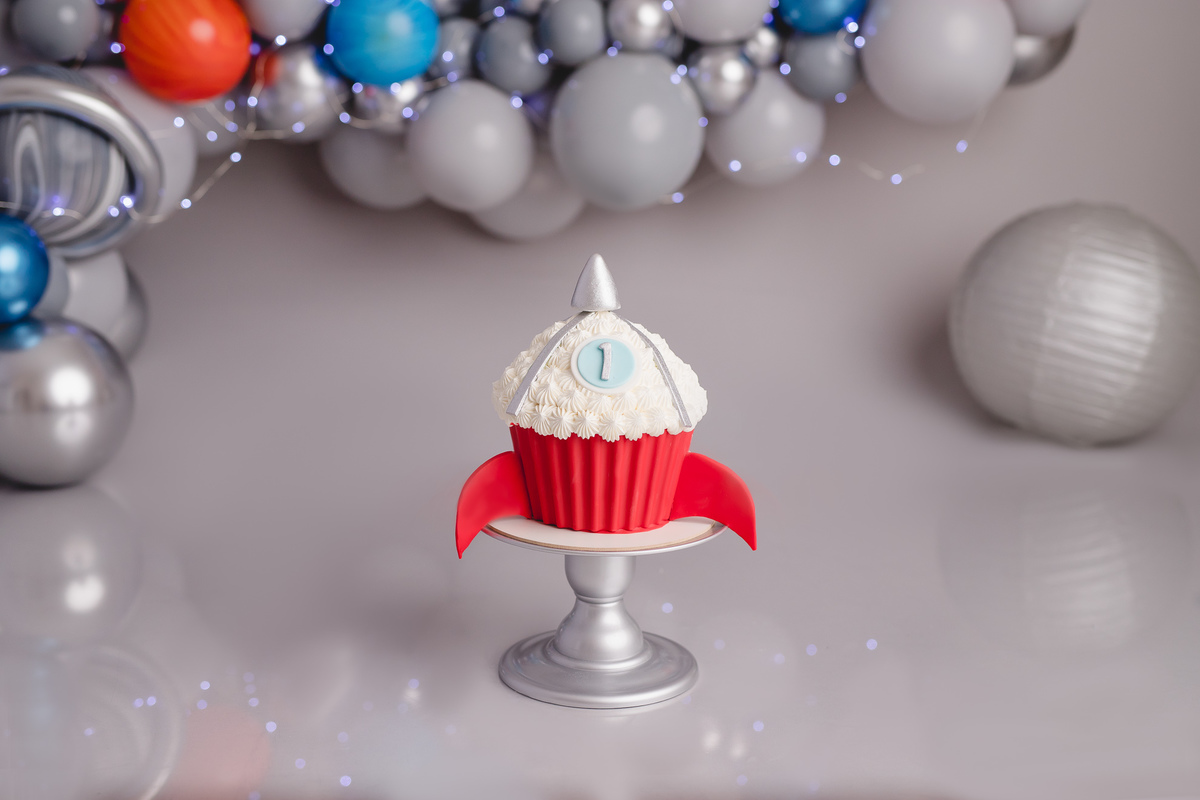 Bebe astronauta no ensaio fotografico de 1 aninho ou primeiro aninho, cenário com fundo cinza e arco de balões imitando planetas e bolo em formato de foguete vermelho com branco em cima de um suporte prata