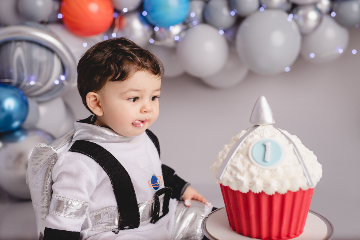 astronauta no ensaio fotografico de 1 aninho ou primeiro aninho cenário cinza e arco de balões imitando planetas bolo em formato de foguete vermelho e branco em um suporte prata, bebê com língua para fora querendo comer o bolo