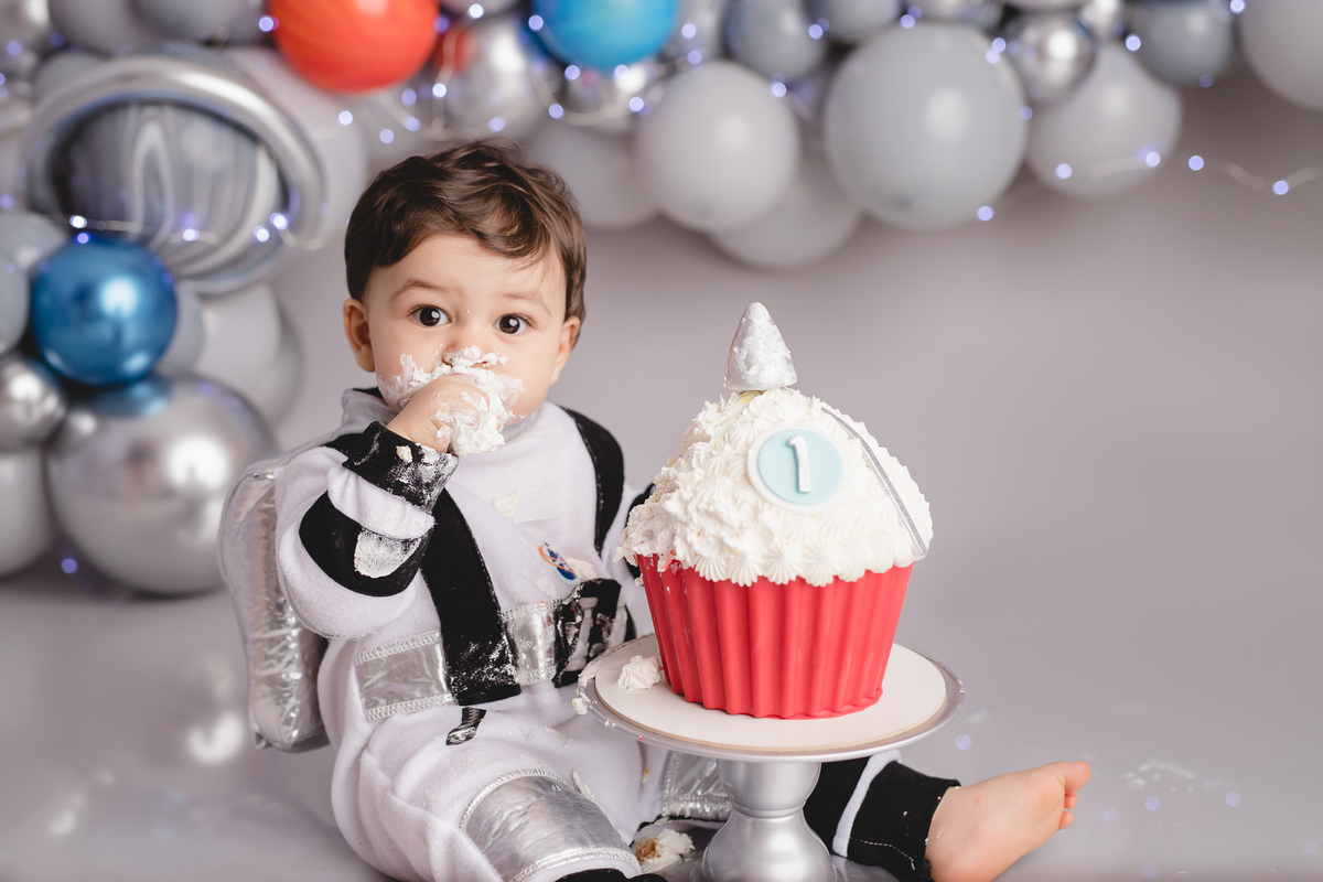 astronauta no ensaio fotografico de 1 aninho ou primeiro aninho cenário cinza e arco de balões imitando planetas bolo em formato de foguete vermelho e branco em um suporte prata, bebê comendo o bolo
