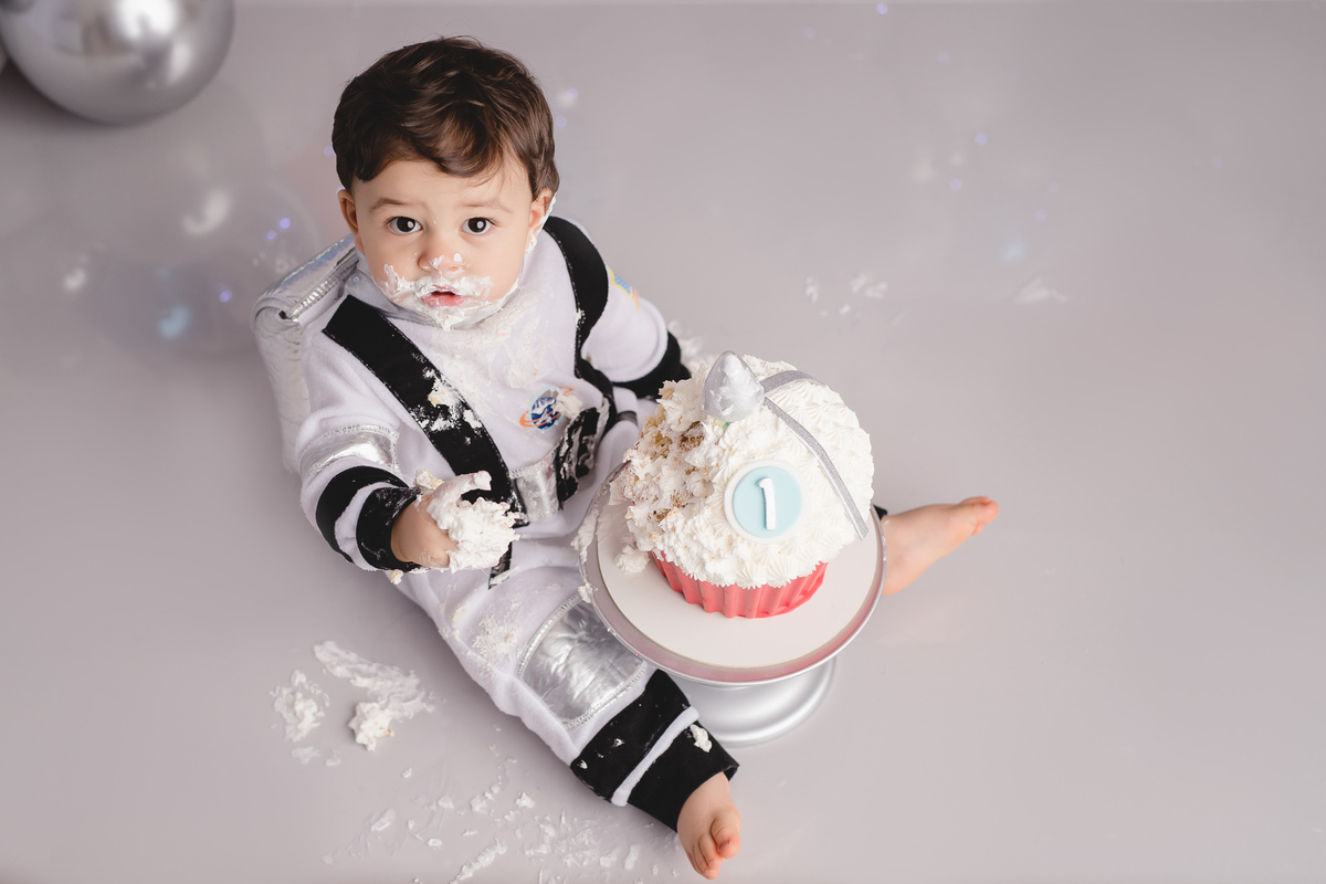 astronauta no ensaio fotografico de 1 aninho ou primeiro aninho cenário cinza e arco de balões imitando planetas bolo em formato de foguete vermelho e branco em um suporte prata, bebê comendo o bolo, imagem vista de cima