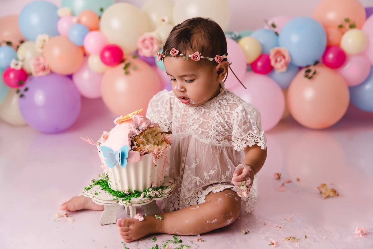 ensaio smash the cake jardim encantado bebe infantil primeiro aninho cadeirinha balões coloridos flores sorrindo roupinha renda e tiara de flores cenário e bolo com borboletas pegando