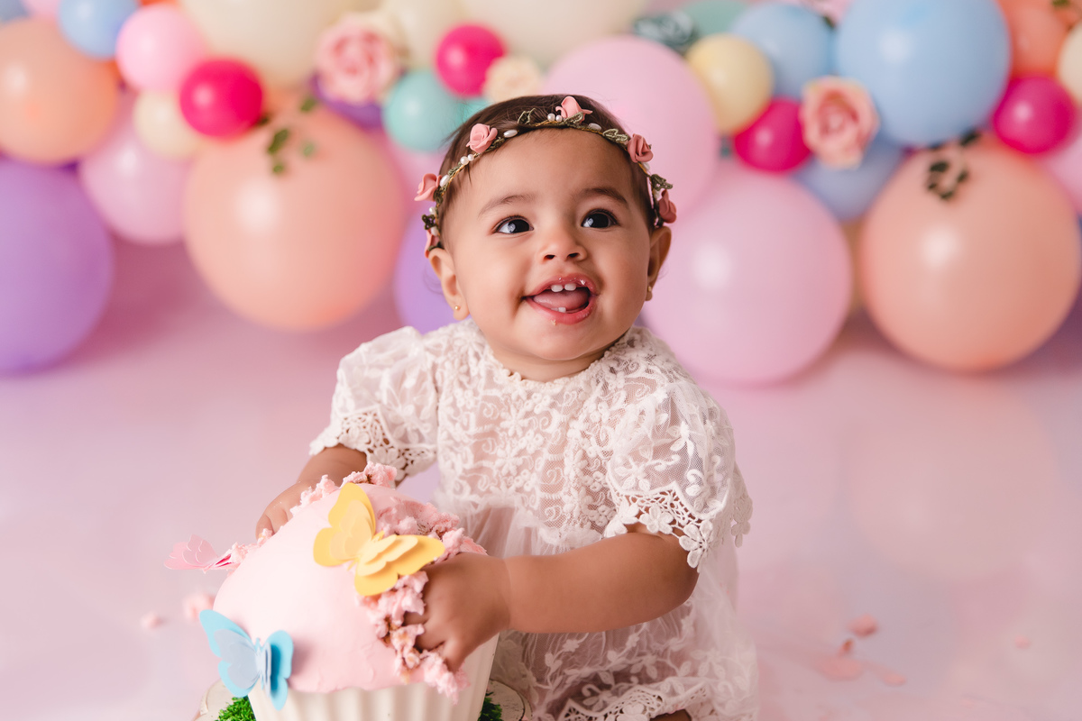 ensaio smash the cake jardim encantado bebe infantil primeiro aninho cadeirinha balões coloridos flores sorrindo roupinha renda e tiara de flores cenário e bolo com borboletas esmagando