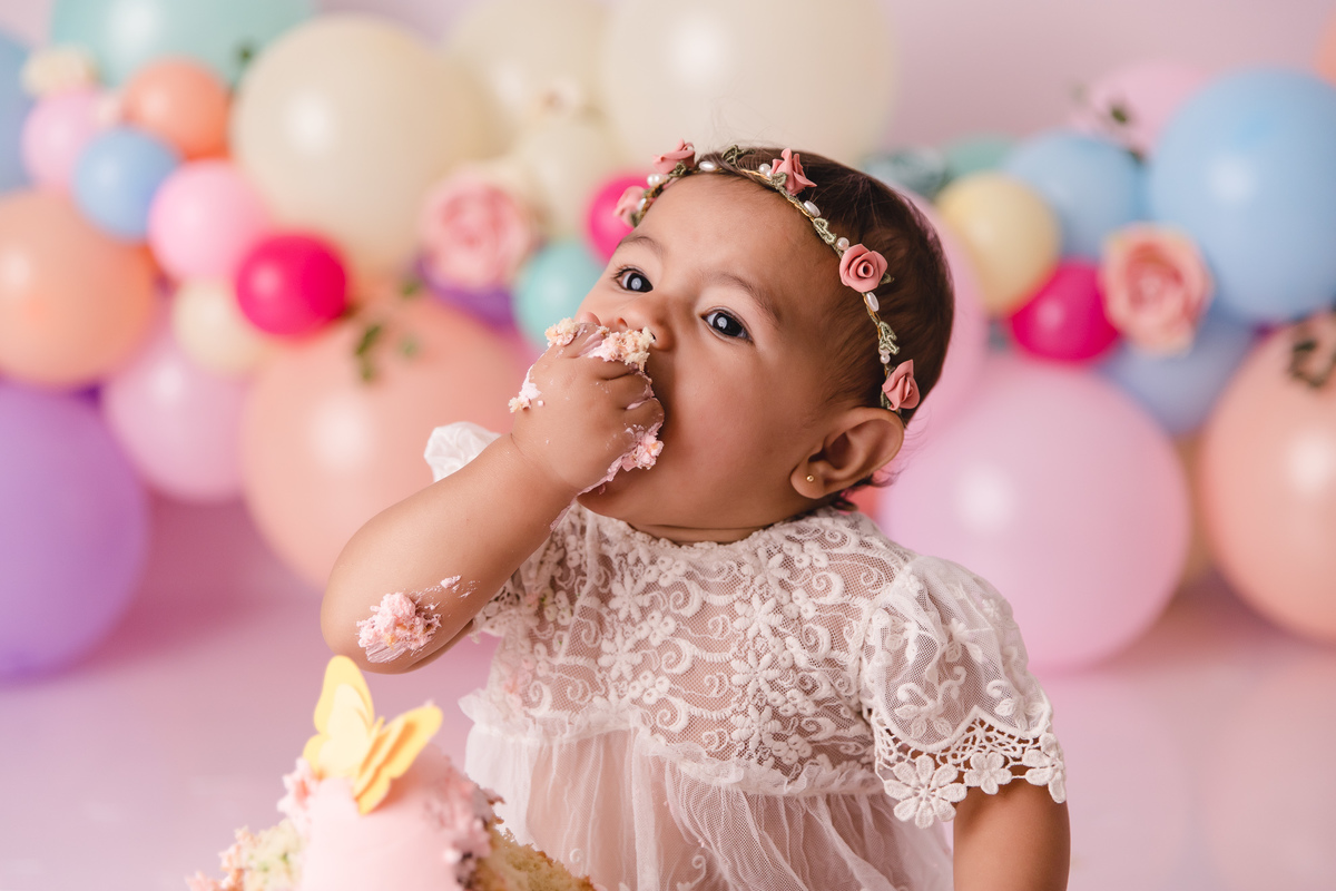 ensaio smash the cake jardim encantado bebe infantil primeiro aninho cadeirinha balões coloridos flores sorrindo roupinha renda e tiara de flores cenário e bolo com borboletas comendo com a mão