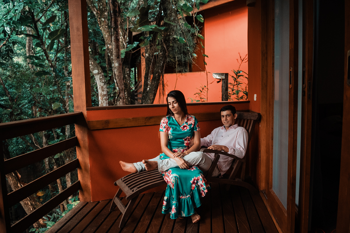 fábio está sentado em uma espreguiçadeira sorrindo enquanto vánubia está sentada entra suas pernas virada para o lado com semblante sereno