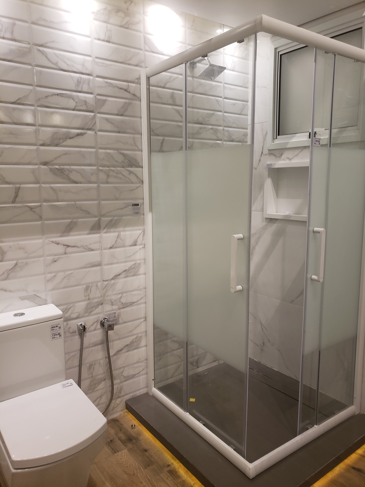 Banheiro branco sem tédio, sofisticado e prático com instalação de nicho no box do banho e ducha higiênica no sanitário.Destaque para a iluminação.