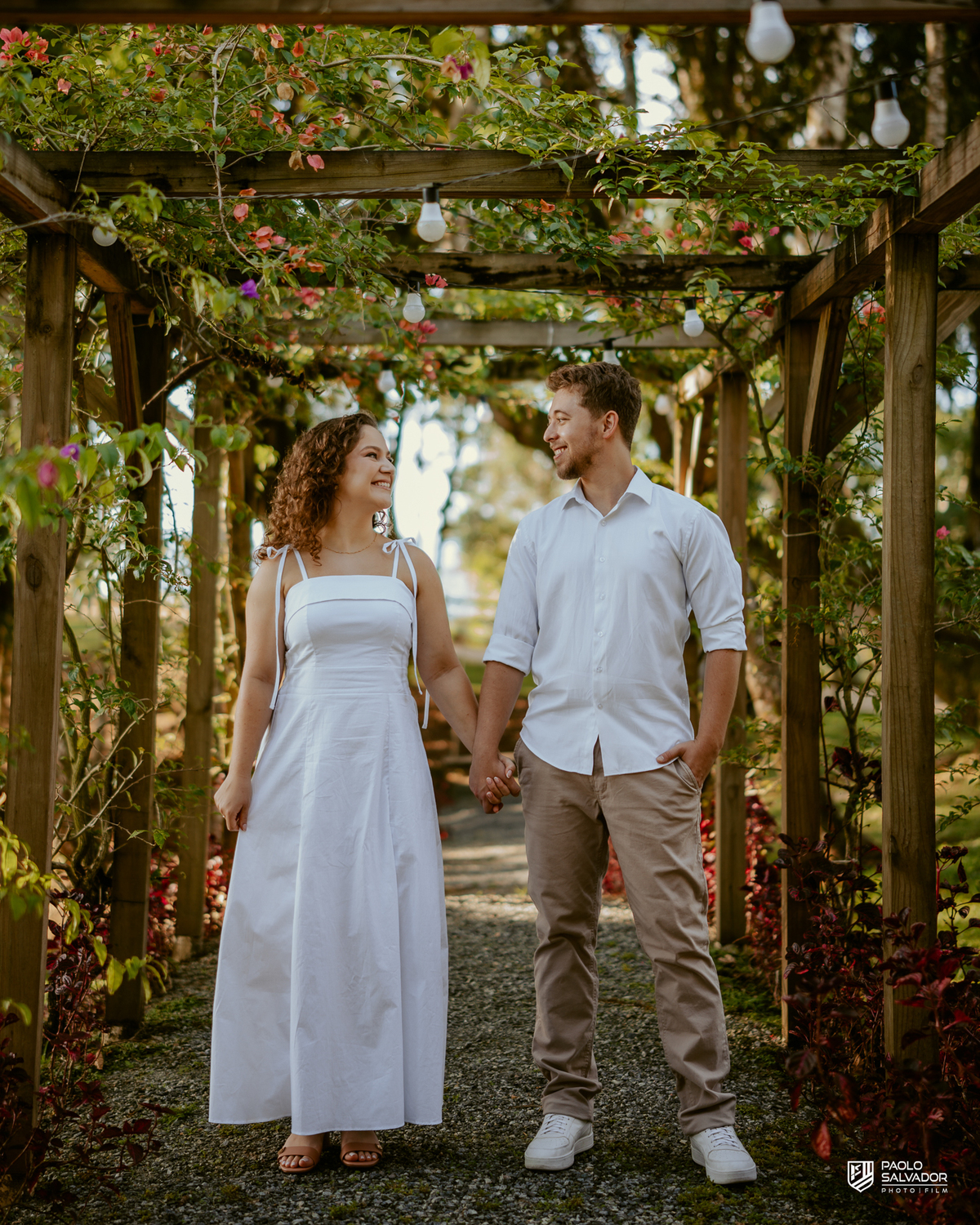 Casal caminhando de mãos dadas em pergolado com flores durante ensaio pré-wedding em Rio dos Cedros, cenário romântico e natural próximo a regiões como Barragem de Palmeiras e Rio Bonito.