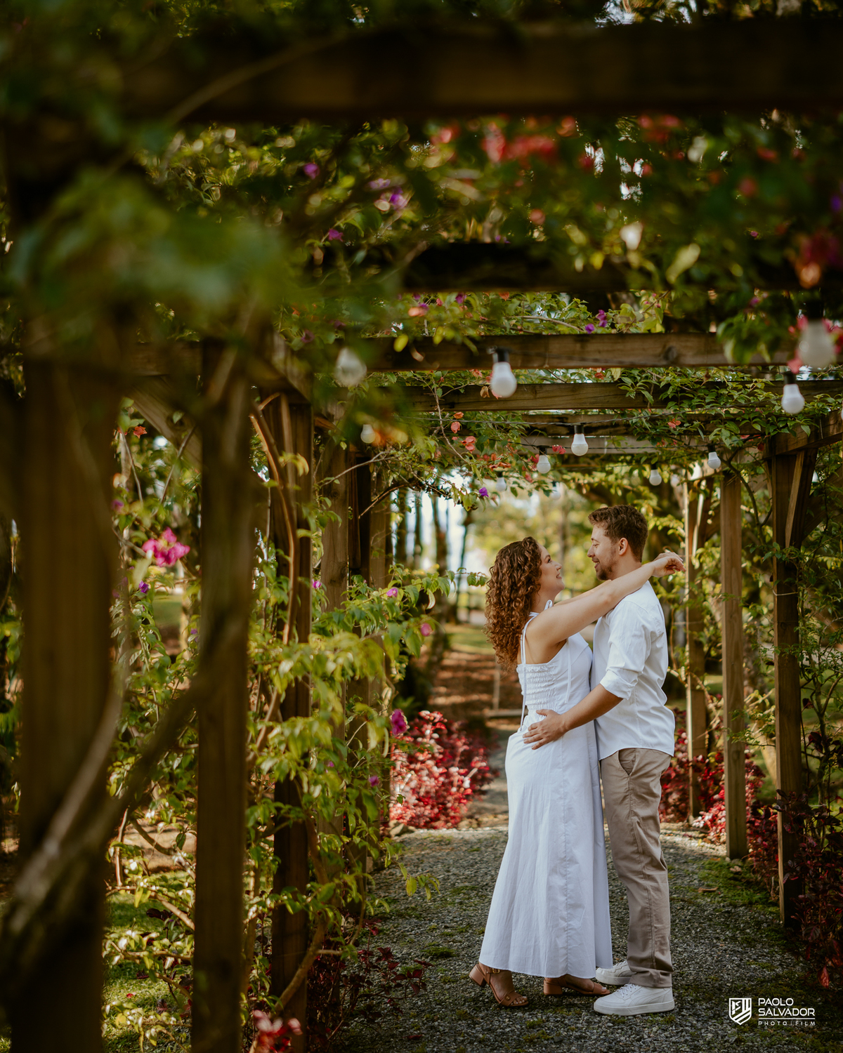 Casal caminhando de mãos dadas em pergolado com flores durante ensaio pré-wedding em Rio dos Cedros, cenário romântico e natural próximo a regiões como Barragem de Palmeiras e Rio Bonito.