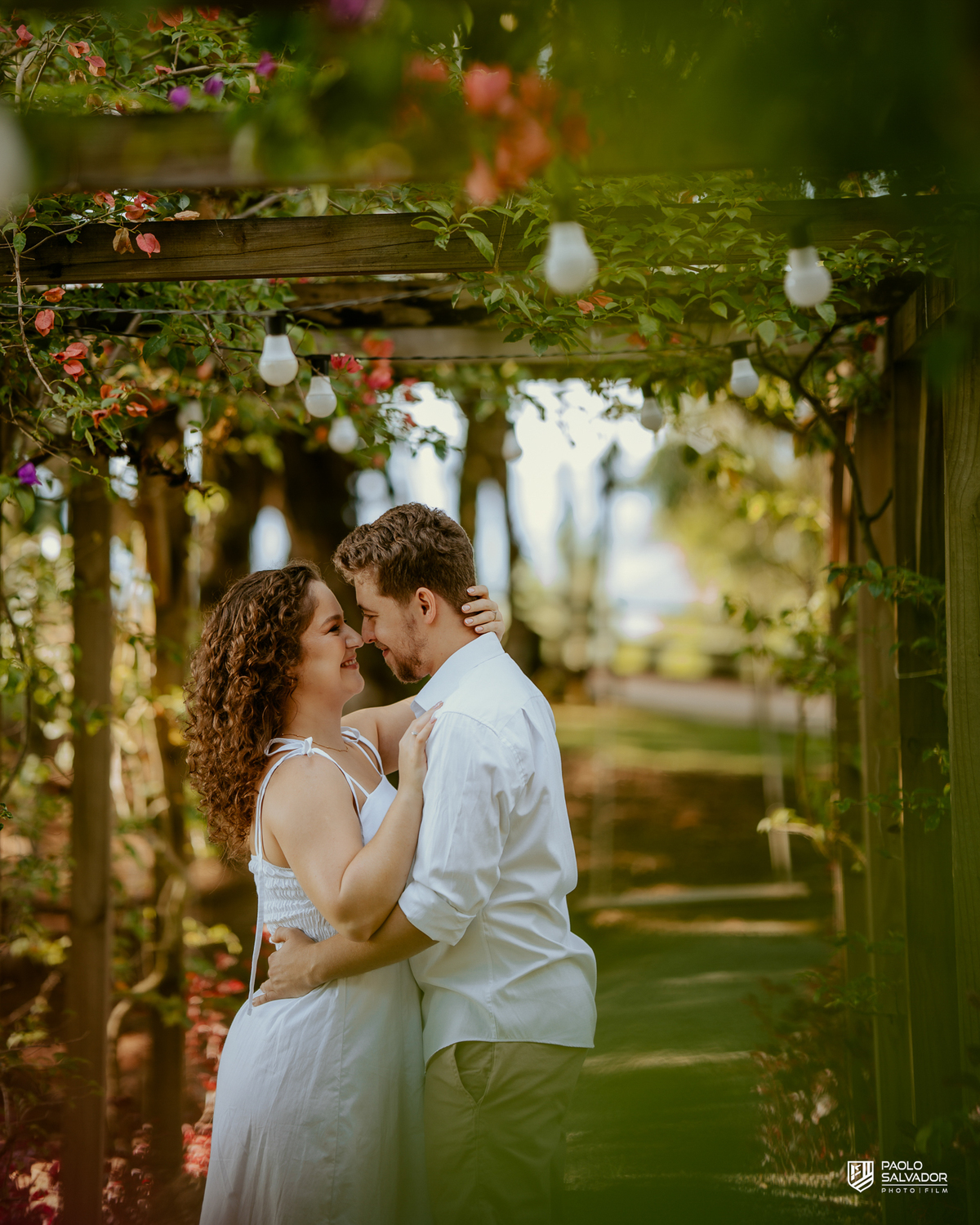 Casal caminhando de mãos dadas em pergolado com flores durante ensaio pré-wedding em Rio dos Cedros, cenário romântico e natural próximo a regiões como Barragem de Palmeiras e Rio Bonito.