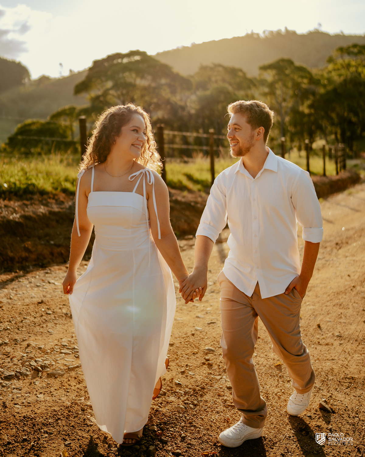 Casal caminhando em estrada rural durante ensaio pré-wedding em Rio dos Cedros, com luz dourada e cenário campestre, inspiração para ensaios na Barragem de Palmeiras, Rio Bonito e região.