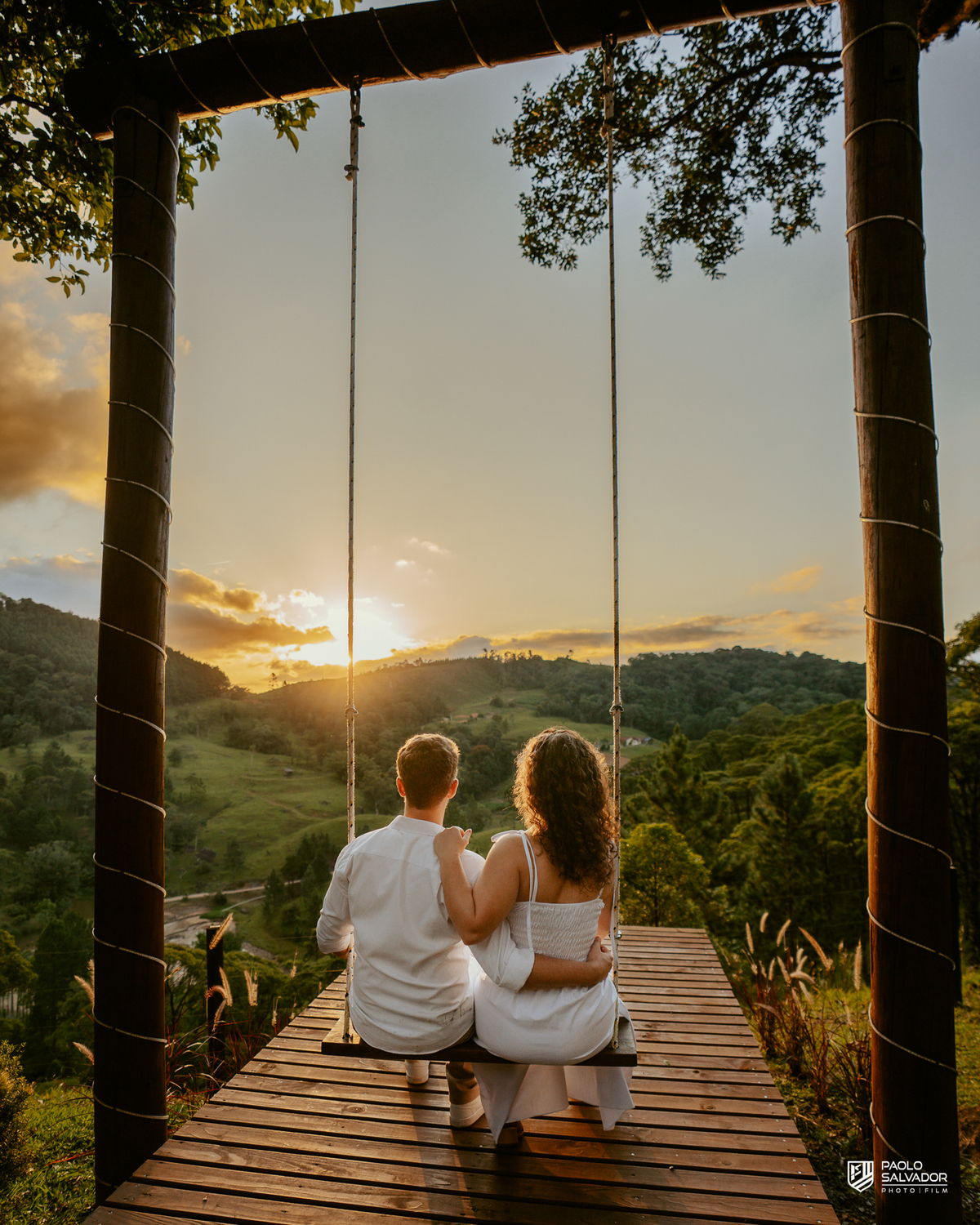 Casal em balanço com vista para montanhas em ensaio pré-wedding em Rio dos Cedros, pôr do sol dourado e paisagem natural, referência para ensaios na Barragem Rio Bonito, Palmeiras e Região dos Lagos.