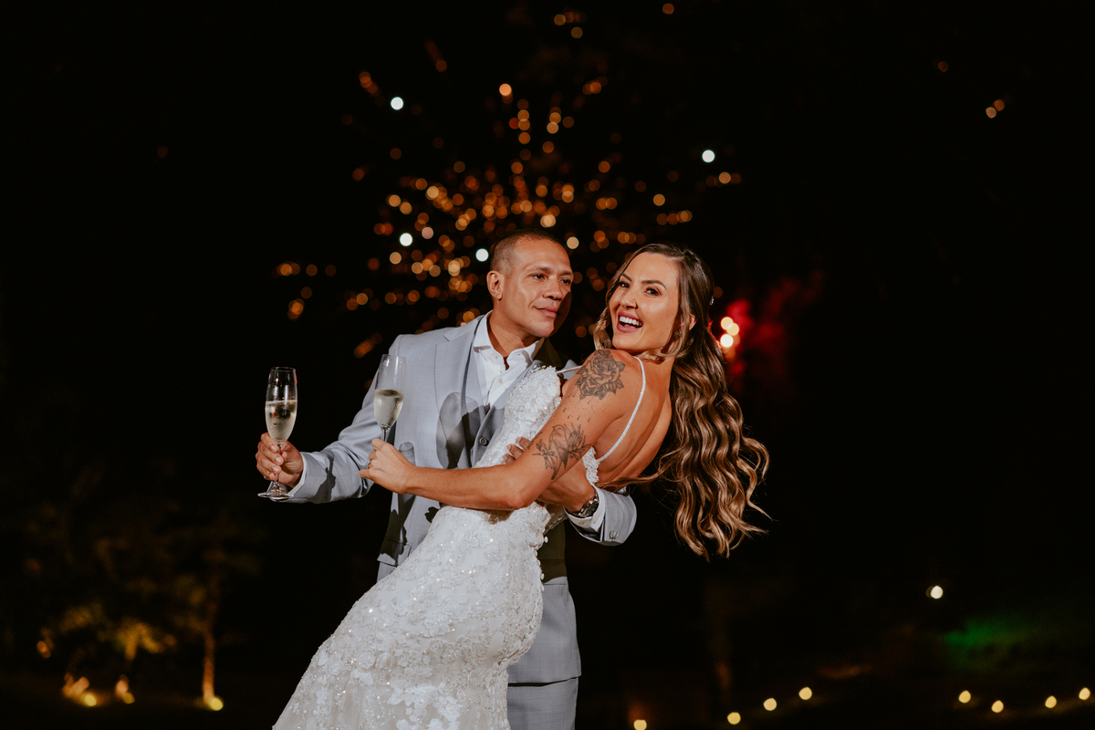 Noivos brindando com espumante durante festa de casamento com fogos ao fundo no Refúgio dos Lagos em Massaranduba, momento marcante e celebração.