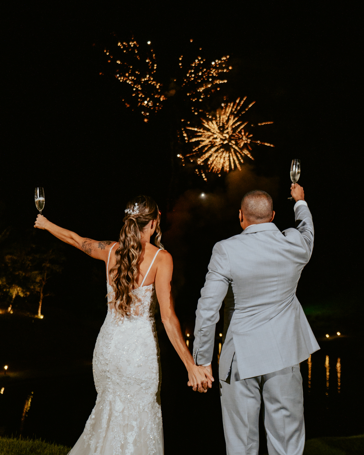 Noivos brindando com espumante durante festa de casamento com fogos ao fundo no Refúgio dos Lagos em Massaranduba, momento marcante e celebração.