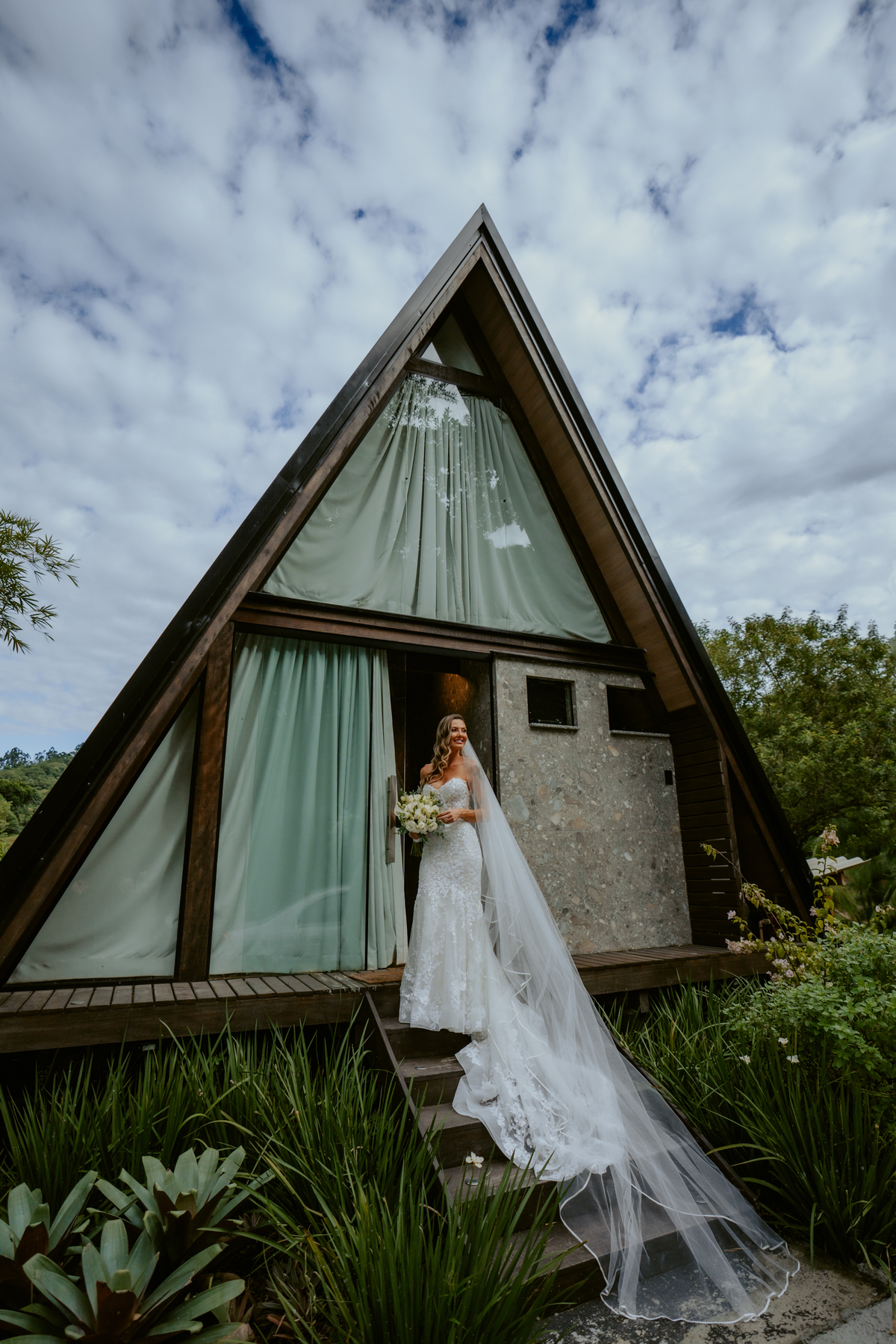 Noiva em frente ao chalé no Refúgio dos Lagos em Massaranduba com véu ao vento, cenário arquitetônico moderno e natureza ao redor, ideal para casamentos ao ar livre.