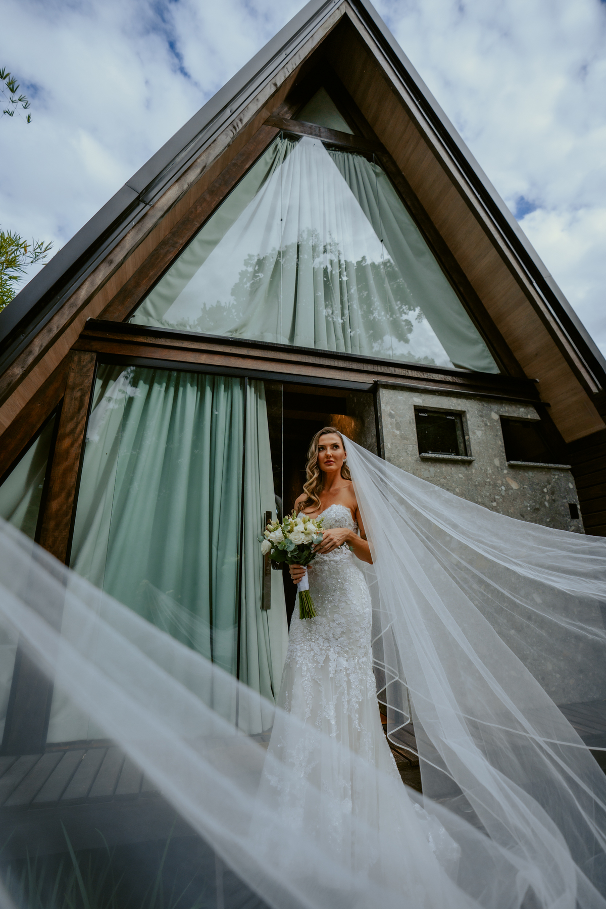 Noiva em frente ao chalé no Refúgio dos Lagos em Massaranduba com véu ao vento, cenário arquitetônico moderno e natureza ao redor, ideal para casamentos ao ar livre.