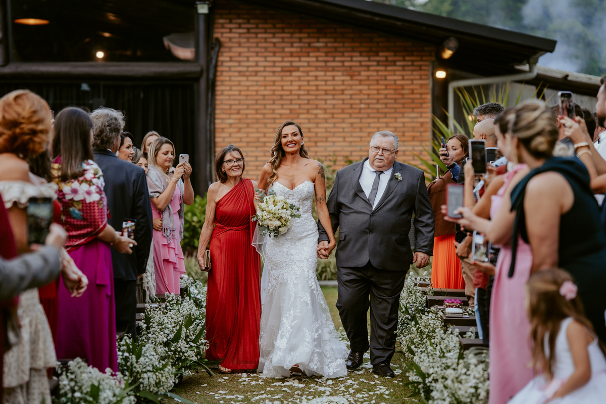 Noiva entrando na cerimônia acompanhada de familiares em casamento ao ar livre no Refúgio dos Lagos em Massaranduba, com convidados emocionados e cenário natural.