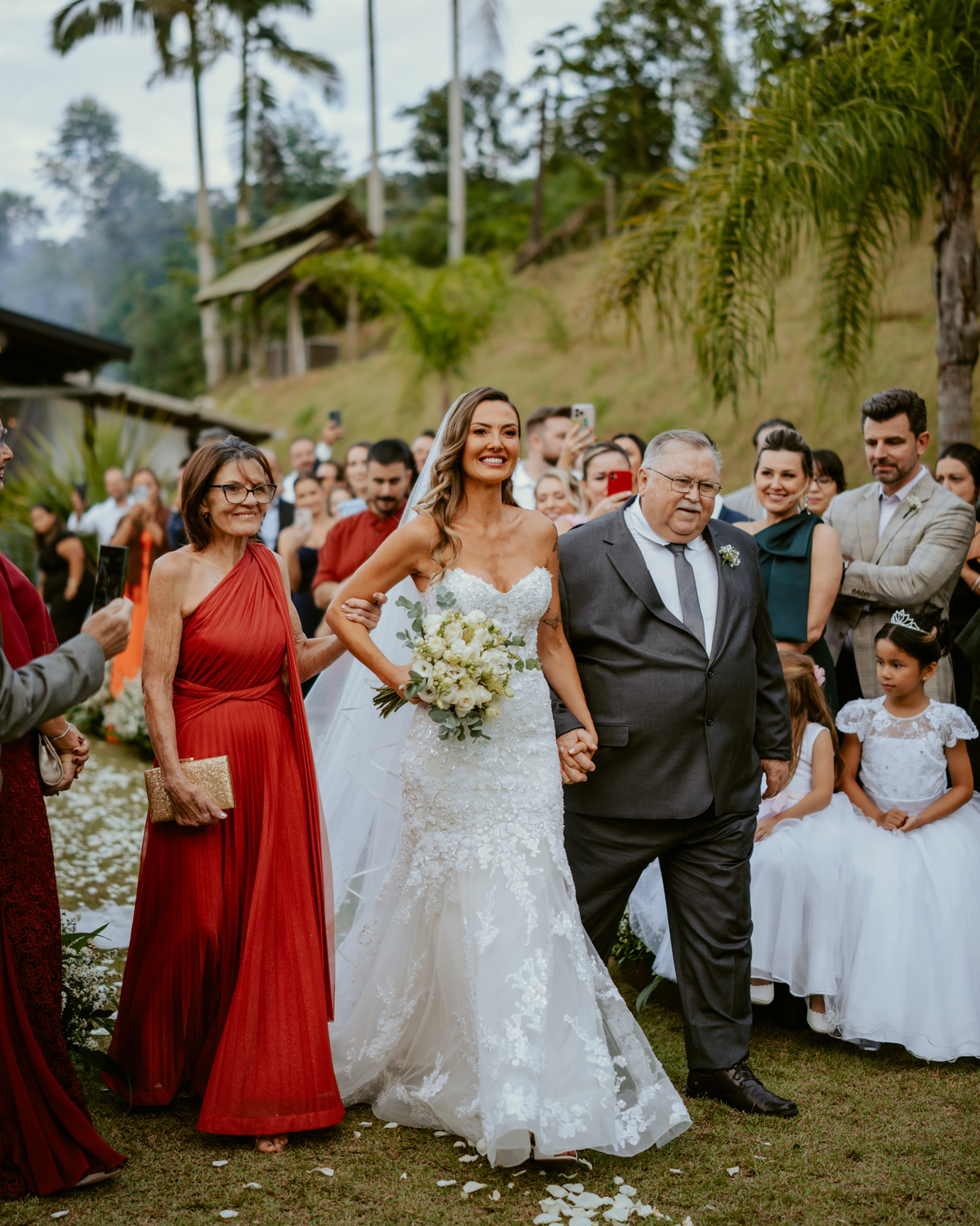 Noiva entrando na cerimônia acompanhada de familiares em casamento ao ar livre no Refúgio dos Lagos em Massaranduba, com convidados emocionados e cenário natural.
