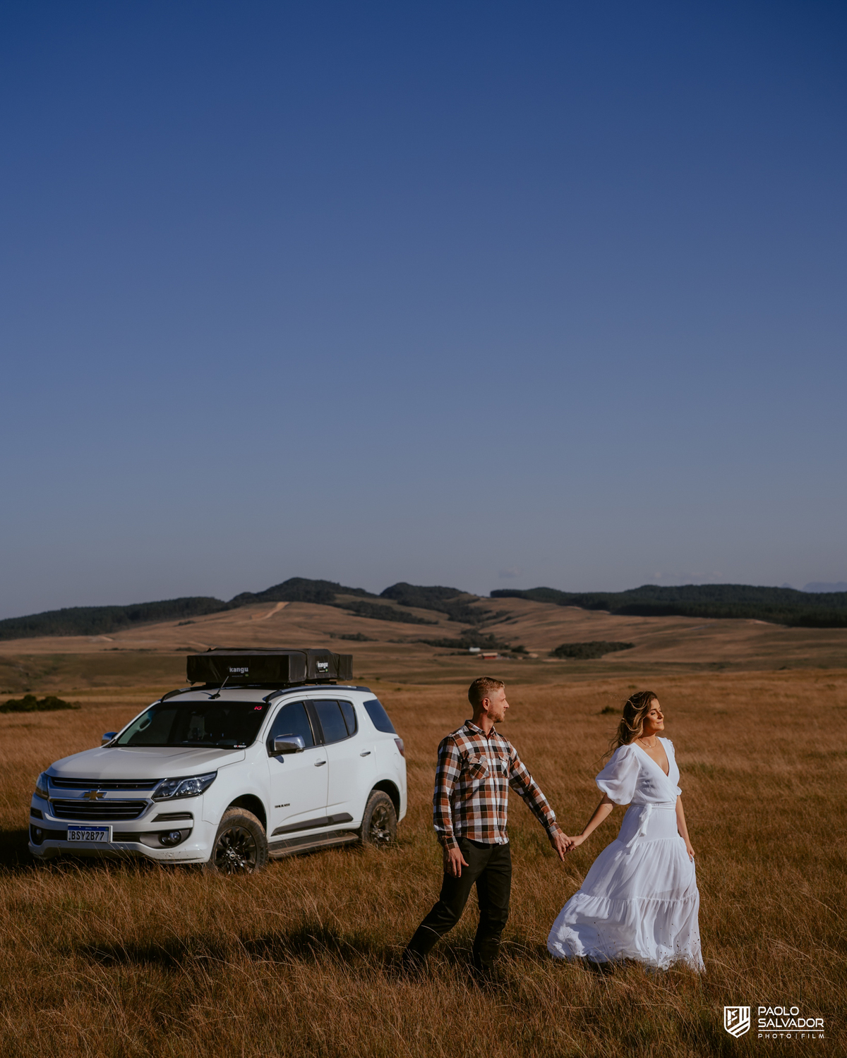 Casal em ensaio pré-wedding em Rancho Queimado no Morro Alto da Boa Vista caminhando de mãos dadas em campo aberto com Trailblazer Chevrolet ao fundo, cenário de montanha e estética natural na Serra Catarinense.
