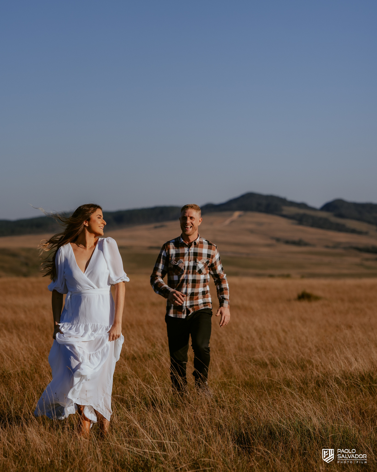 Casal caminhando em campo aberto durante ensaio pré-wedding na Serra Catarinense em Rancho Queimado, com pôr do sol e montanhas ao fundo, estilo romântico e natural.
