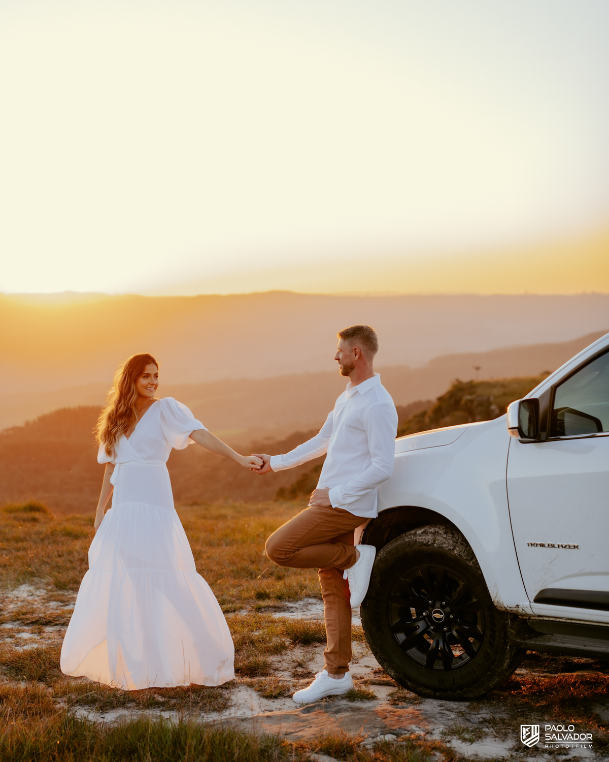 Casal sentado na traseira da Trailblazer com barraca de camping durante ensaio pré-wedding em Rancho Queimado, com pôr do sol e paisagem cinematográfica.