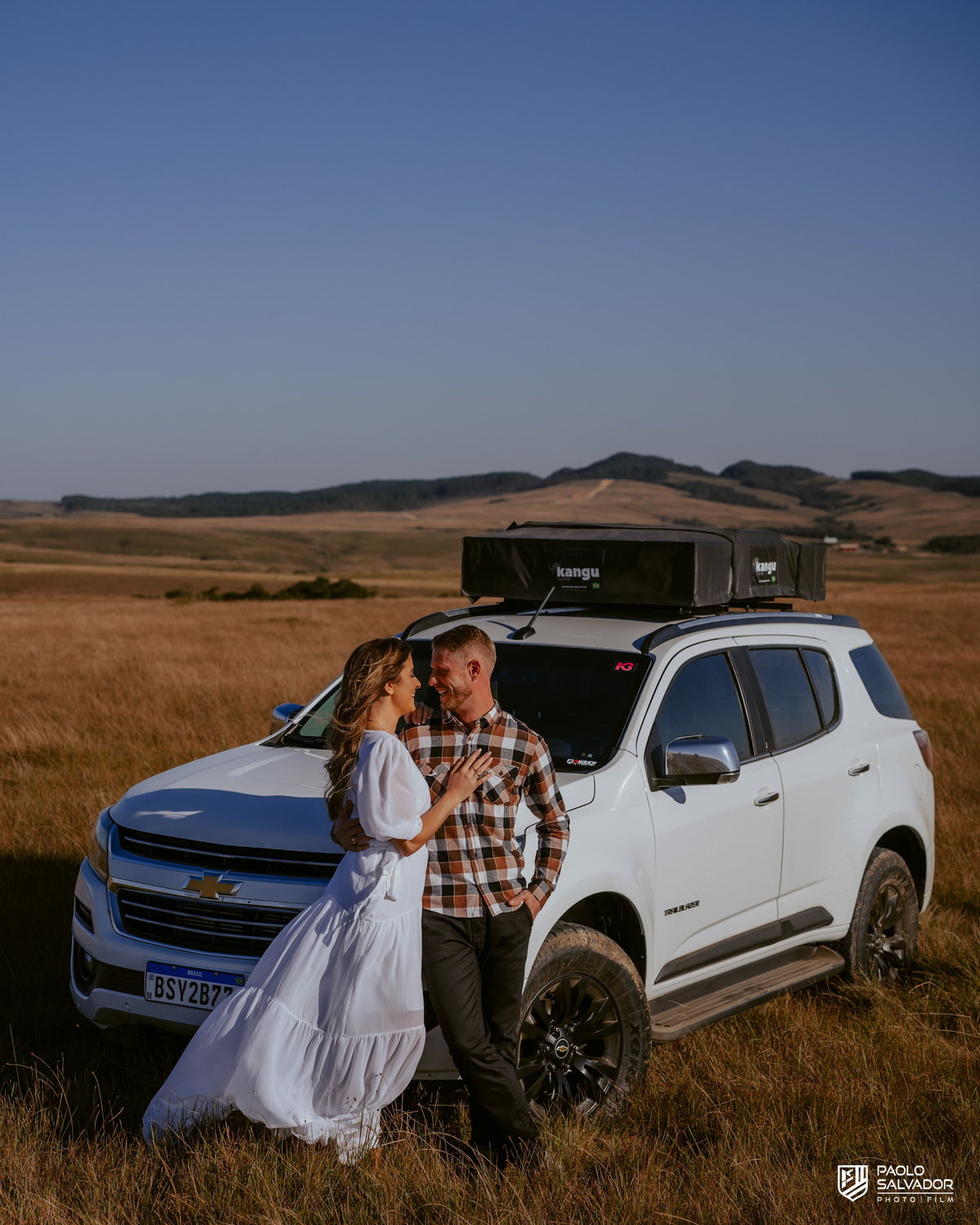 Casal em ensaio pré-wedding em Rancho Queimado no Morro Alto da Boa Vista caminhando de mãos dadas em campo aberto com Trailblazer Chevrolet ao fundo, cenário de montanha e estética natural na Serra Catarinense.