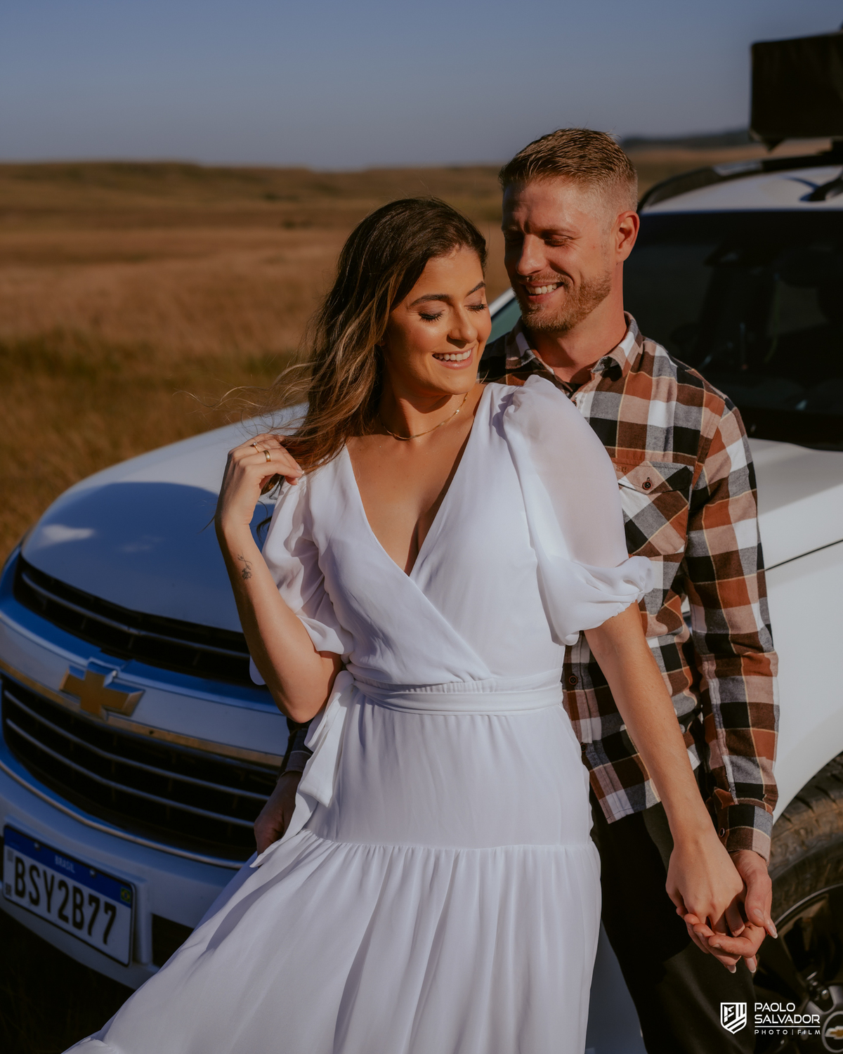 Casal Tainá e Leandro abraçados em frente à caminhonete Trailblazer durante ensaio pré-wedding em Rancho Queimado, com luz dourada e paisagem de campo típica da Serra Catarinense.