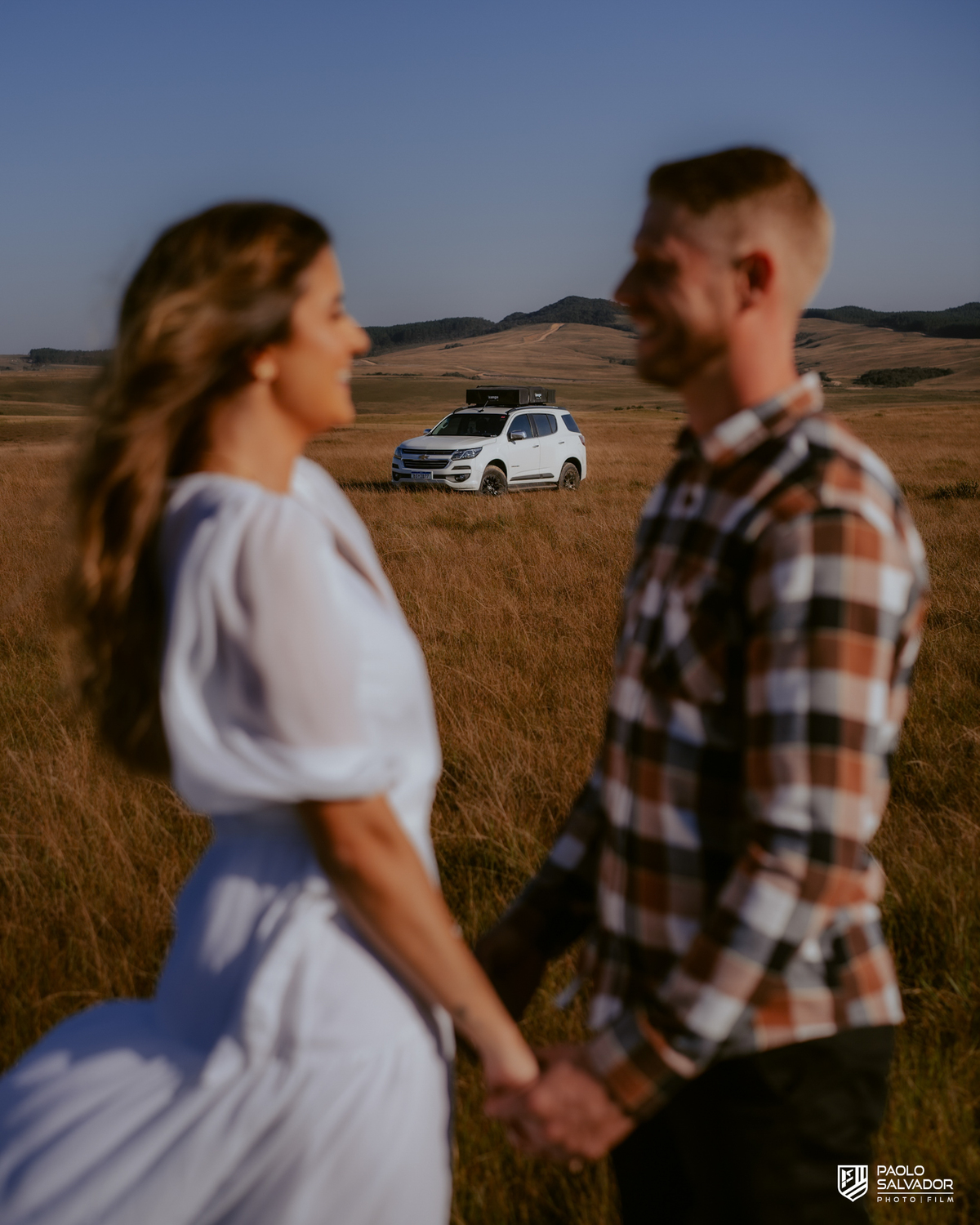 Casal em ensaio pré-wedding em Rancho Queimado no Morro Alto da Boa Vista caminhando de mãos dadas em campo aberto com Trailblazer Chevrolet ao fundo, cenário de montanha e estética natural na Serra Catarinense.