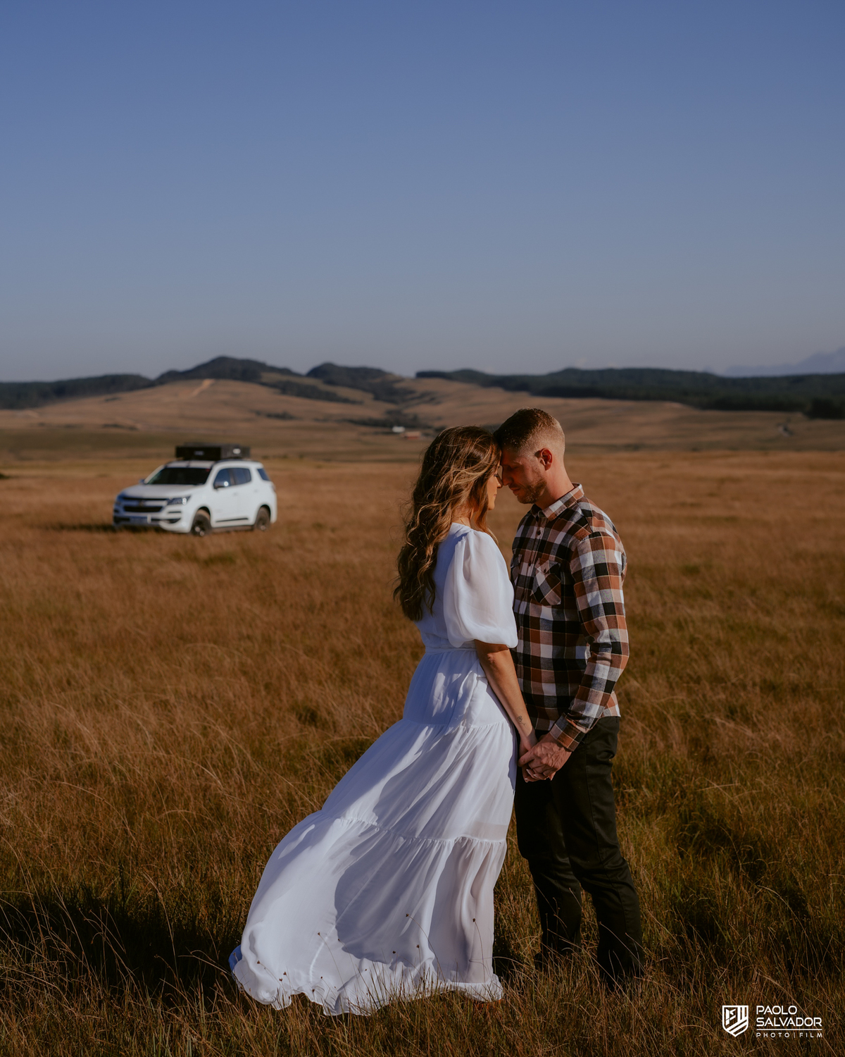 Casal em ensaio pré-wedding em Rancho Queimado no Morro Alto da Boa Vista caminhando de mãos dadas em campo aberto com Trailblazer Chevrolet ao fundo, cenário de montanha e estética natural na Serra Catarinense.