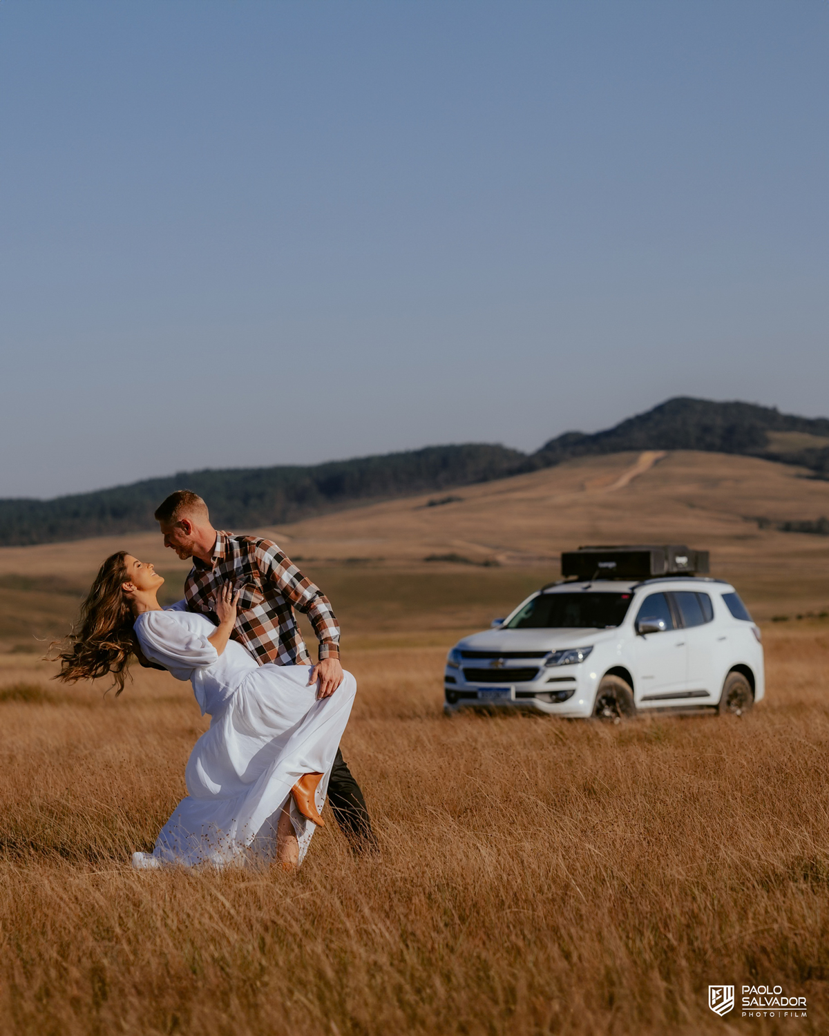 Casal Tainá e Leandro abraçados em frente à caminhonete Trailblazer durante ensaio pré-wedding em Rancho Queimado, com luz dourada e paisagem de campo típica da Serra Catarinense.