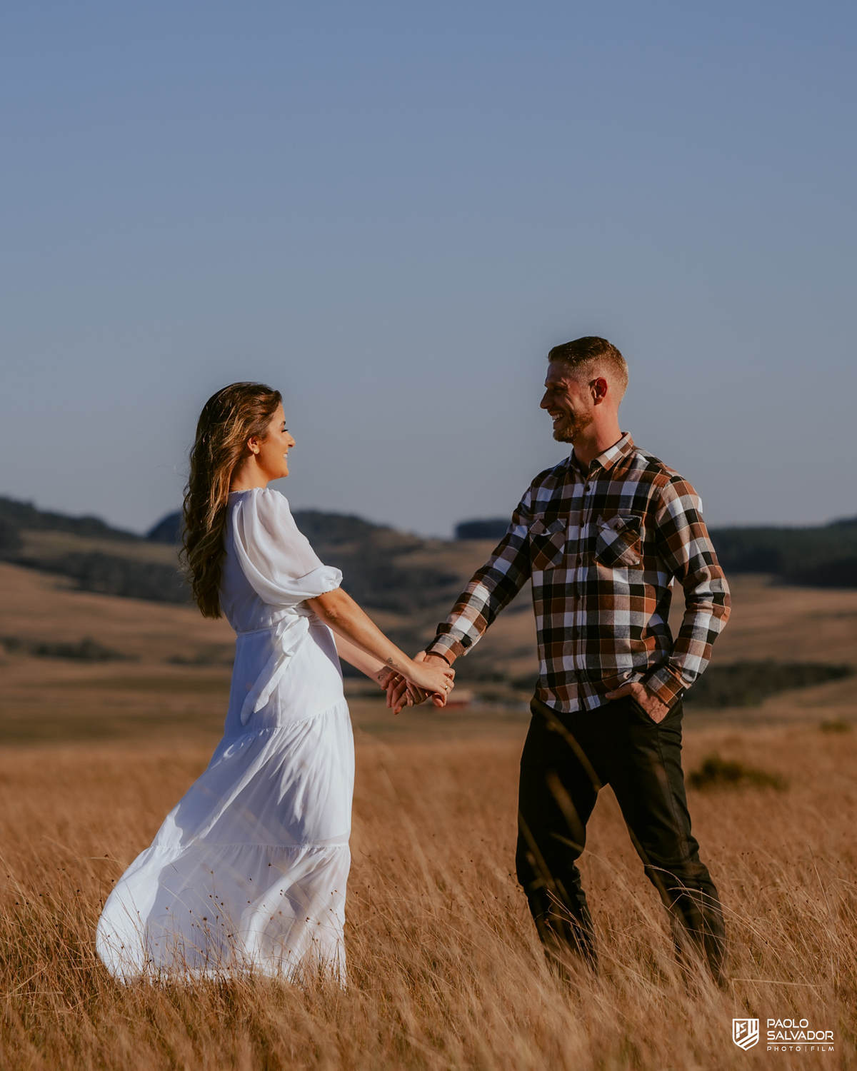 Casal caminhando em campo aberto durante ensaio pré-wedding na Serra Catarinense em Rancho Queimado, com pôr do sol e montanhas ao fundo, estilo romântico e natural.
