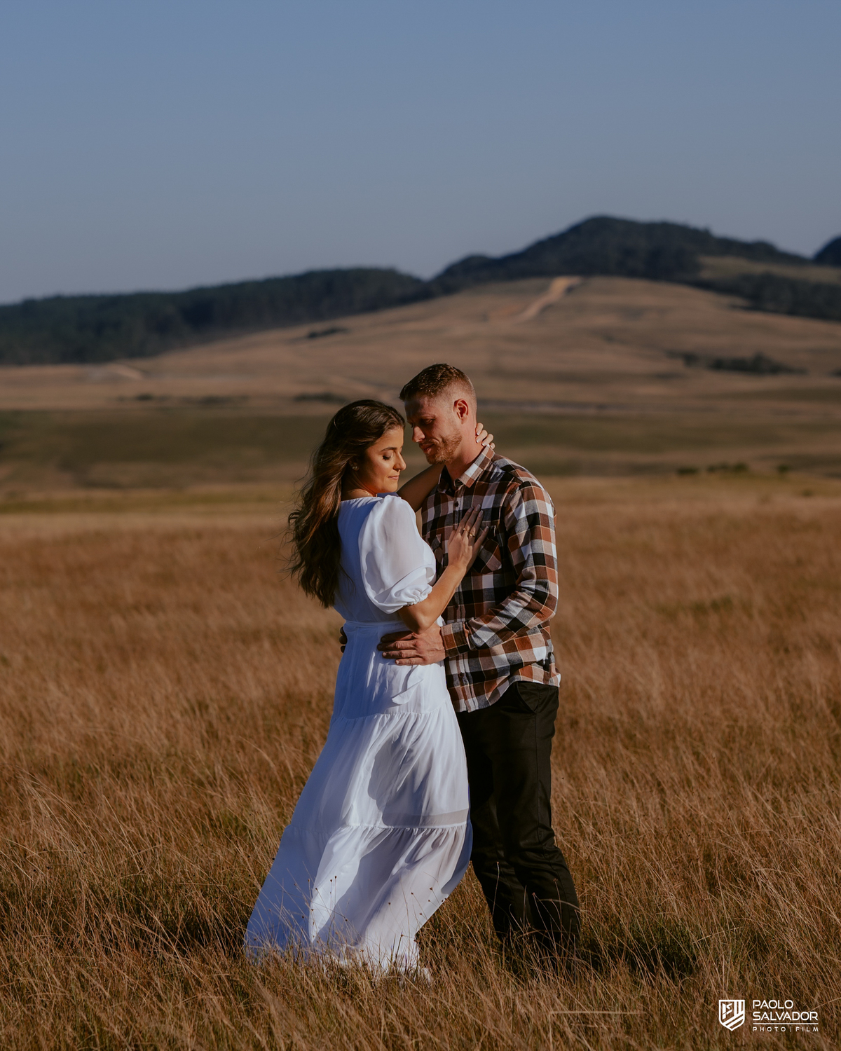 Casal caminhando em campo aberto durante ensaio pré-wedding na Serra Catarinense em Rancho Queimado, com pôr do sol e montanhas ao fundo, estilo romântico e natural.
