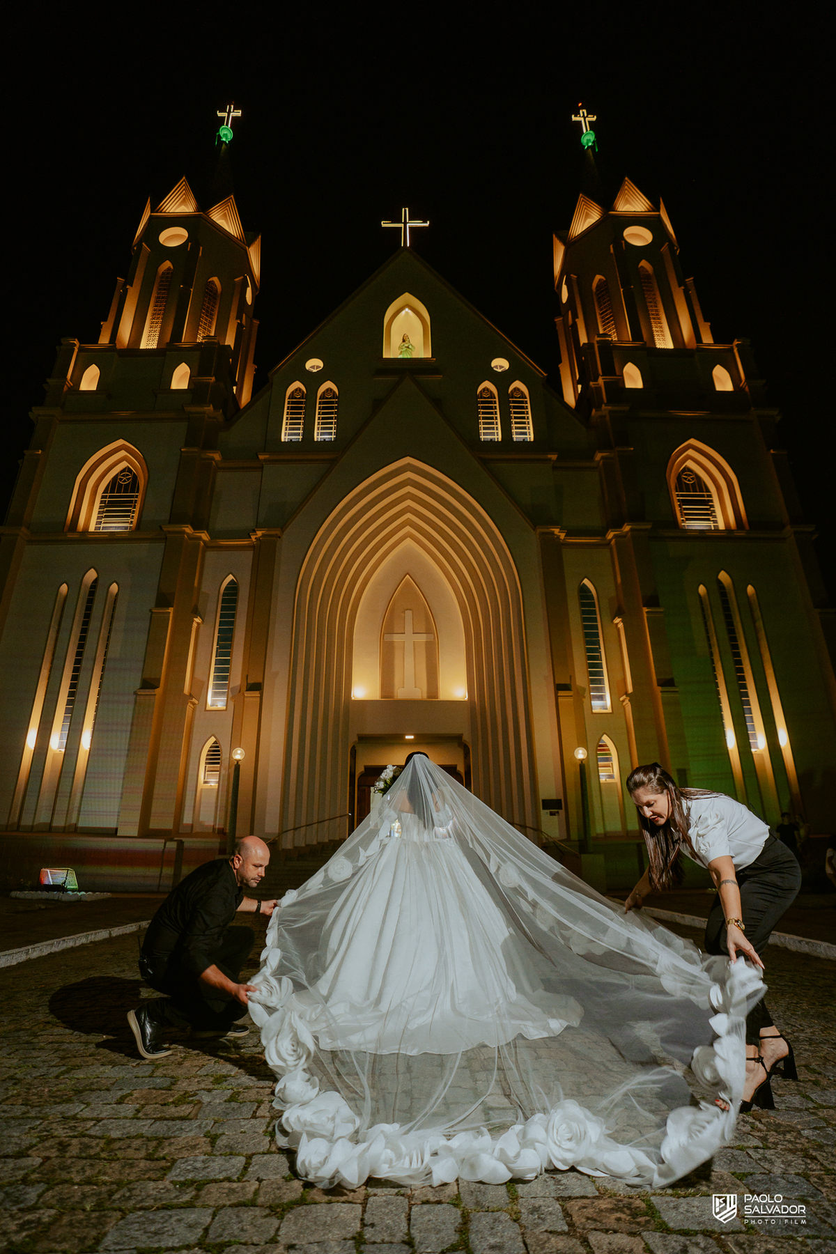 Noiva com véu sendo ajustado pela equipe em frente à Igreja Santa Inês em Indaial à noite, preparação para ensaio pós-cerimônia, fotografia de casamento noturno