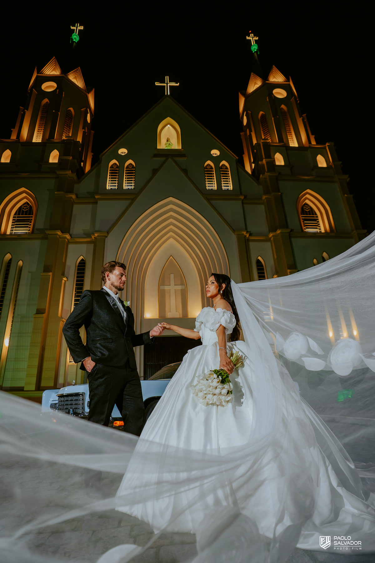 Retrato do casal com carro clássico na frente da Igreja Santa Inês em Indaial à noite, fotografia elegante e romântica de casamento SC