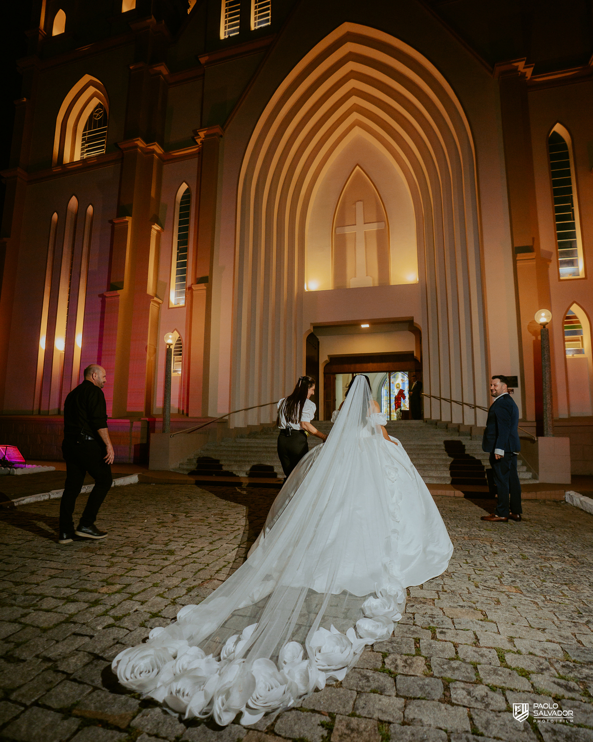 Noiva com véu sendo ajustado pela equipe em frente à Igreja Santa Inês em Indaial à noite, preparação para ensaio pós-cerimônia, fotografia de casamento noturno