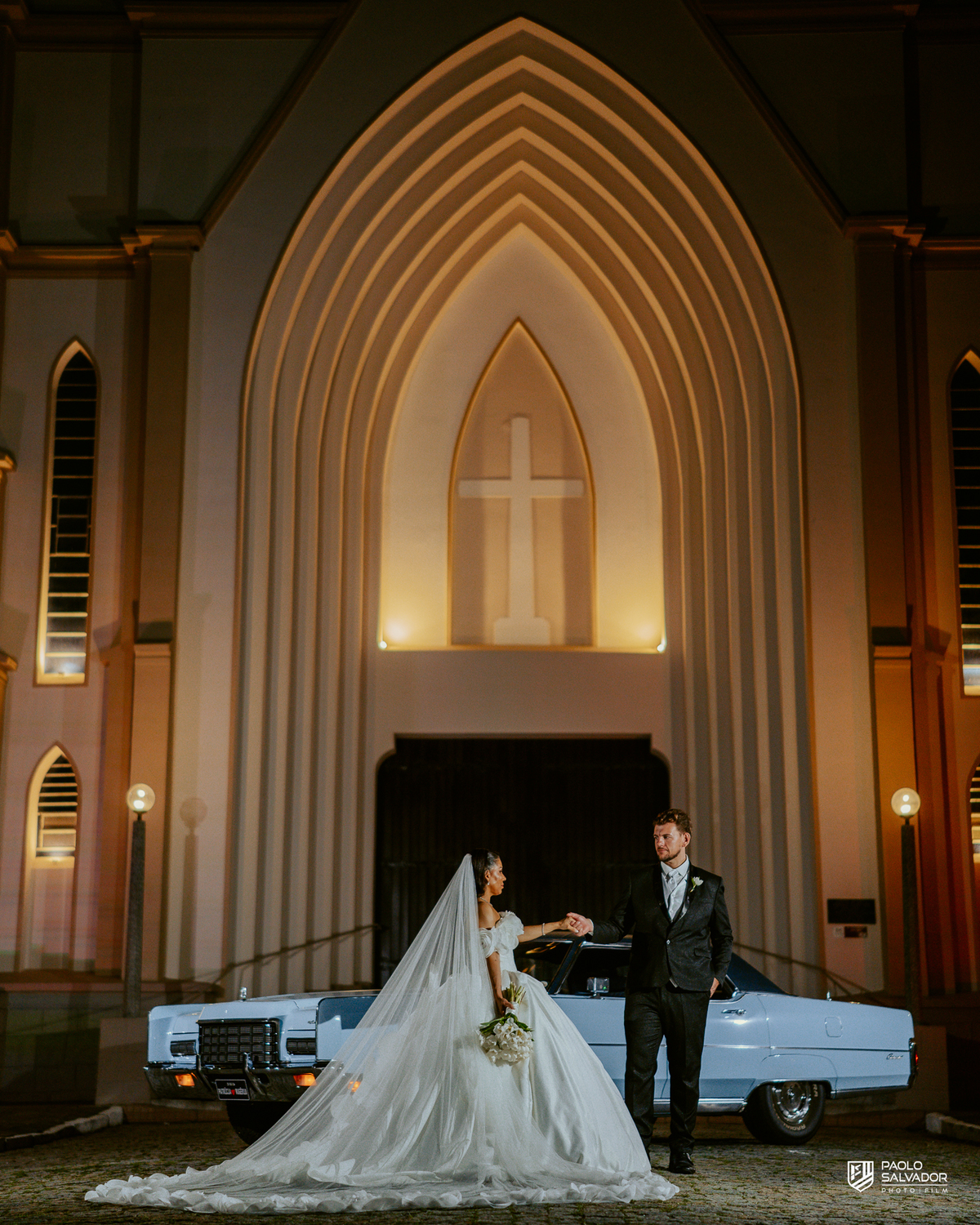 Retrato do casal com carro clássico na frente da Igreja Santa Inês em Indaial à noite, fotografia elegante e romântica de casamento SC