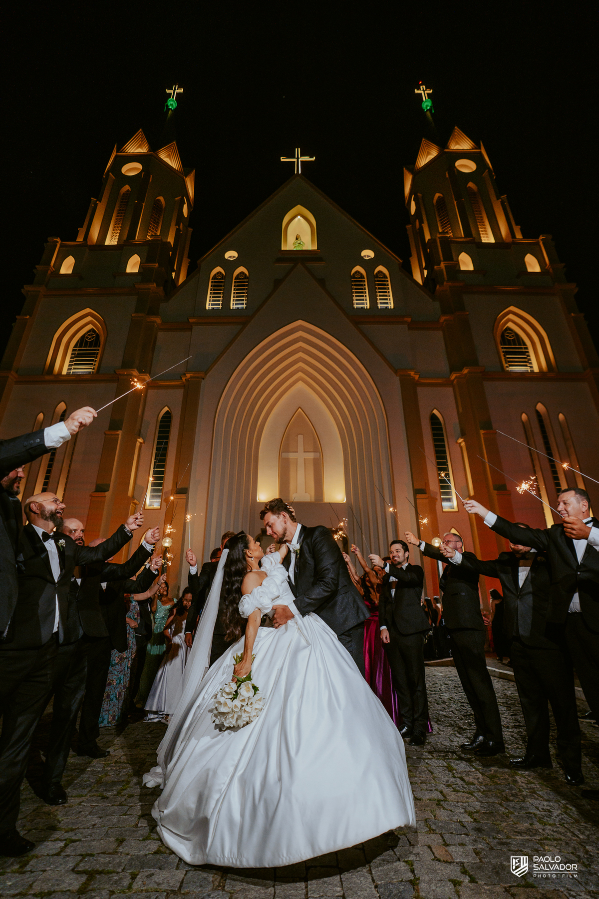 Saída dos noivos com sparkles em frente à Igreja Santa Inês em Indaial, beijo do casal, fotografia de casamento noturno cinematográfico