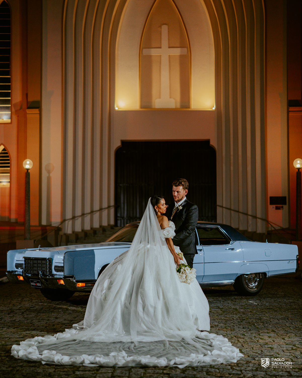 Retrato do casal com carro clássico na frente da Igreja Santa Inês em Indaial à noite, fotografia elegante e romântica de casamento SC
